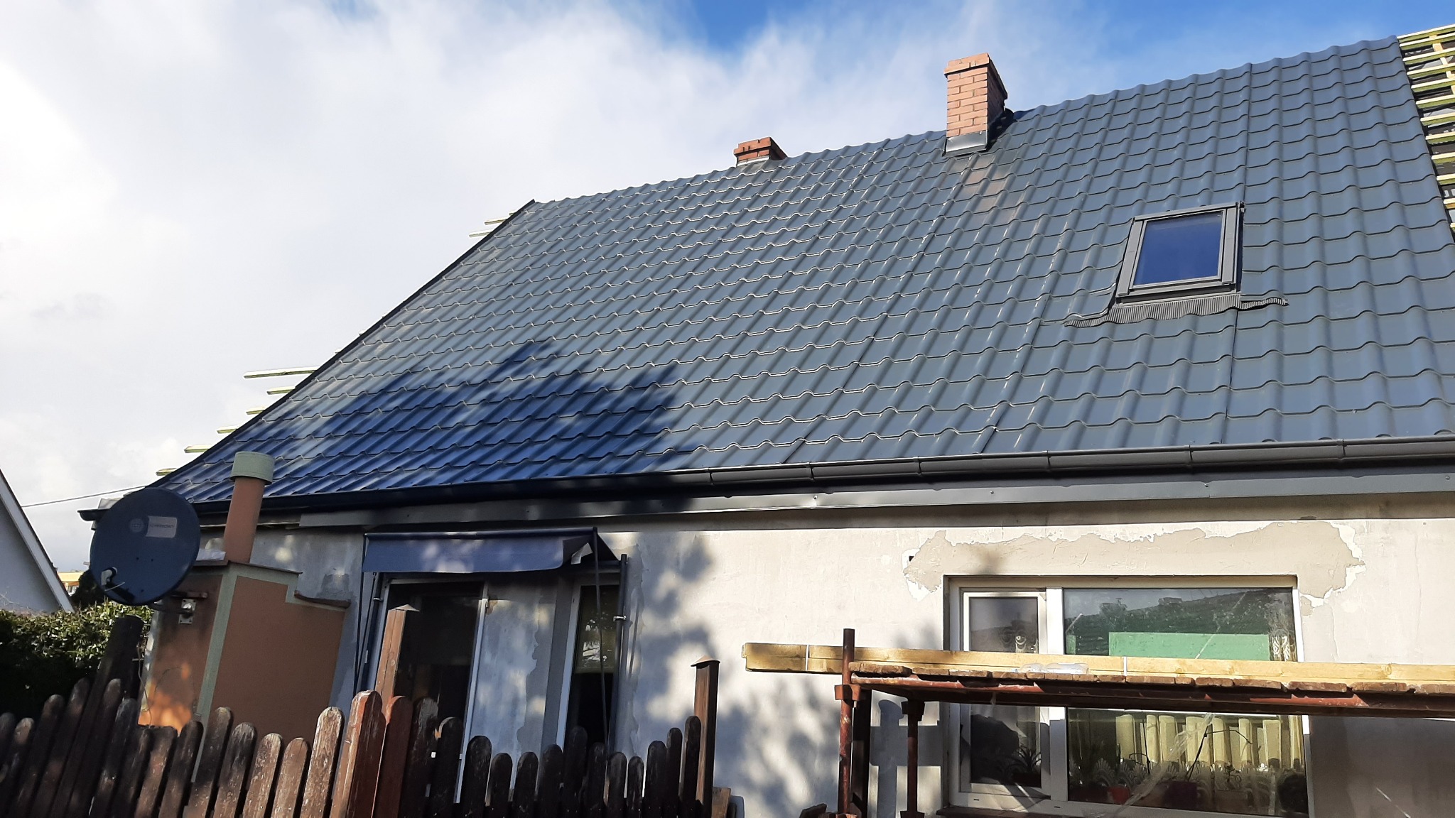 Dom z nowym, grafitowym dachem z dachówki metalowej, widoczne kominy z cegły, okno dachowe i zamontowane rynny.