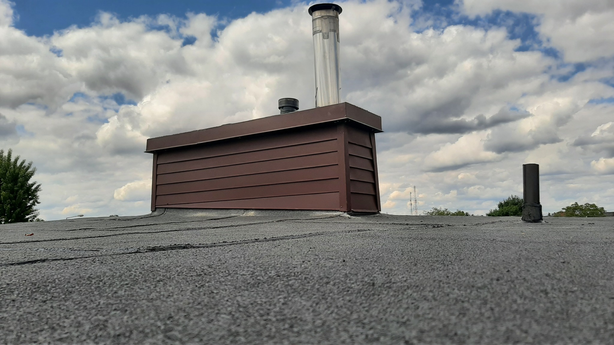 Szczegółowe ujęcie dachu pokrytego papą z brązową obudową komina i metalowym kominem wentylacyjnym na tle pochmurnego nieba.