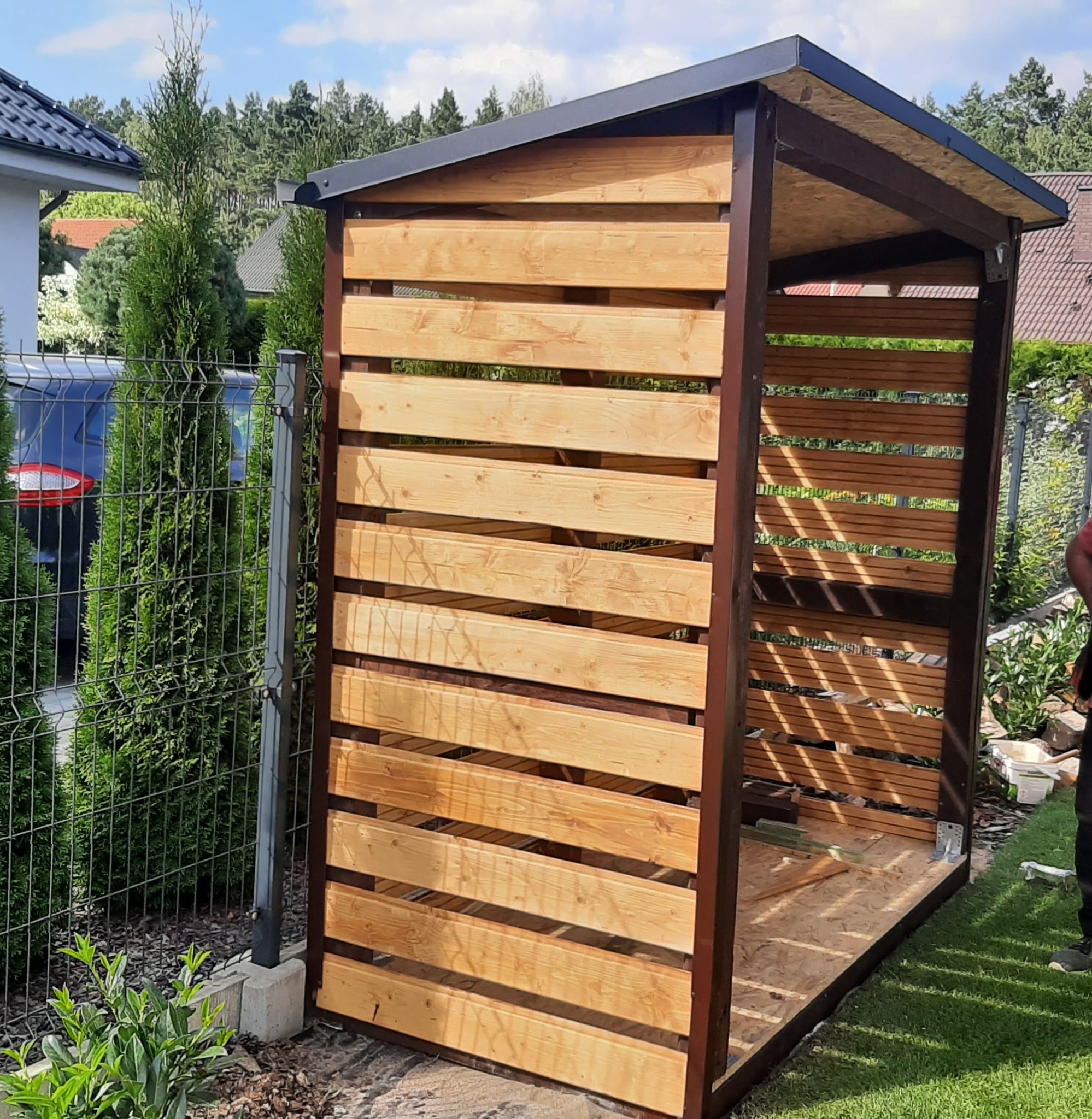 Drewniana konstrukcja altany z poziomymi listwami, brązowe metalowe słupki, widok z boku na tle zielonego trawnika i ogrodzenia z siatki.