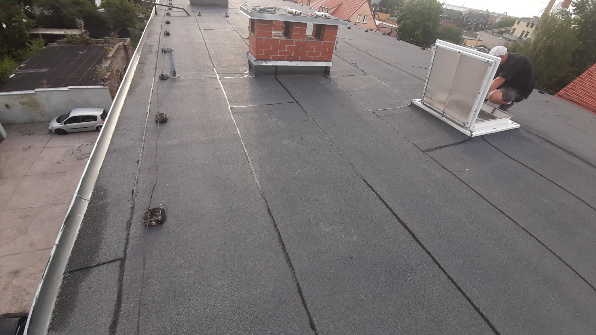 Pracownik w trakcie montażu okna dachowego na płaskim dachu pokrytym papą, widoczne narzędzia i materiały przy kominie z cegły.