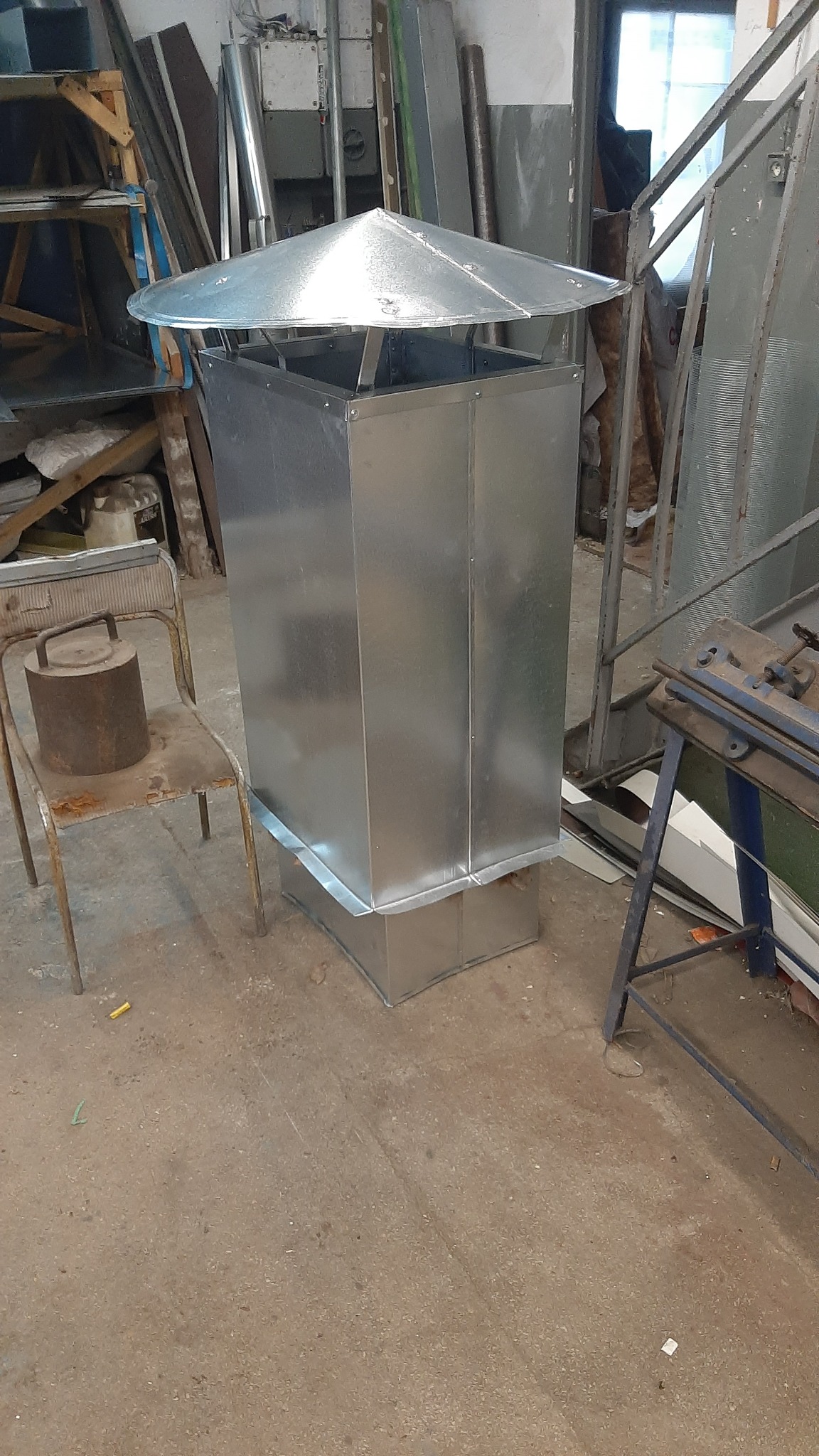 Nowy, błyszczący, kwadratowy komin wentylacyjny z metalu z daszkiem, ustawiony w warsztacie obok starego krzesła z obciążnikiem i narzędzi do obróbki metali.