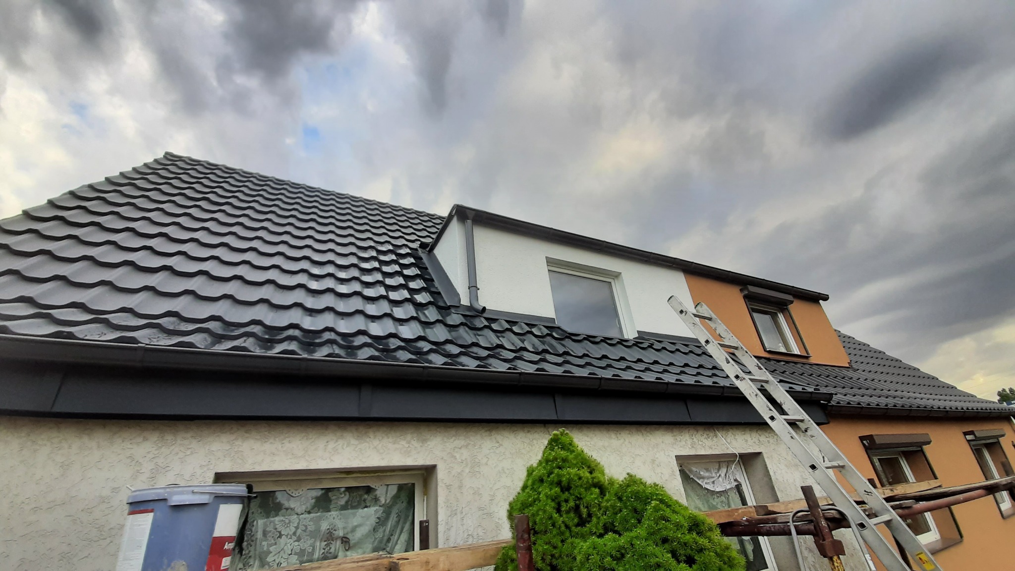 Nowy, błyszczący, grafitowy dach z blachodachówki trapezowej na domu z oknami dachowymi, widok z dołu na tle pochmurnego nieba, drabina oparta o ścianę.