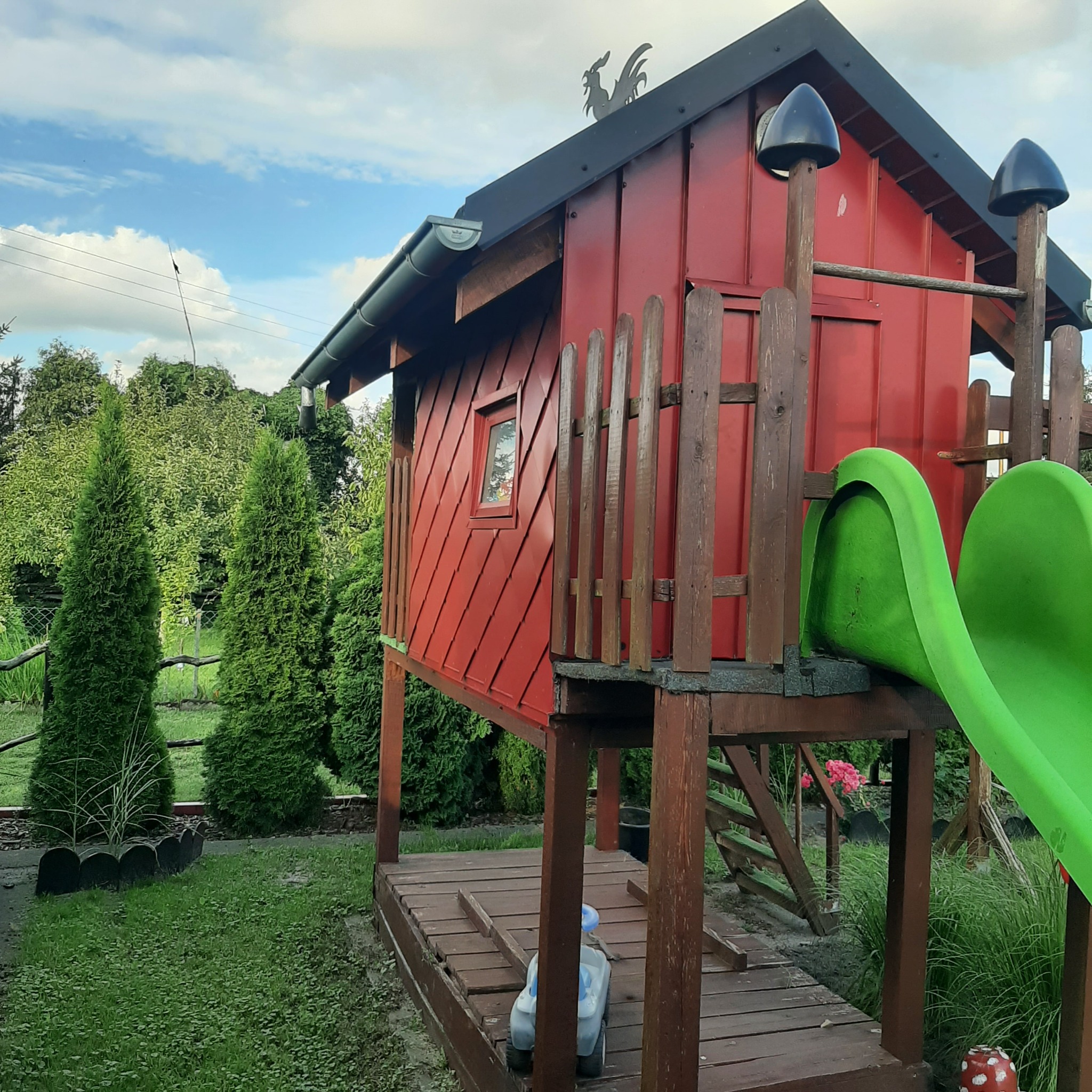 Czerwony domek zabaw dla dzieci z zieloną zjeżdżalnią, metalowym kogutem na dachu i drewnianymi elementami wykończeniowymi, otoczony zielenią ogrodu.