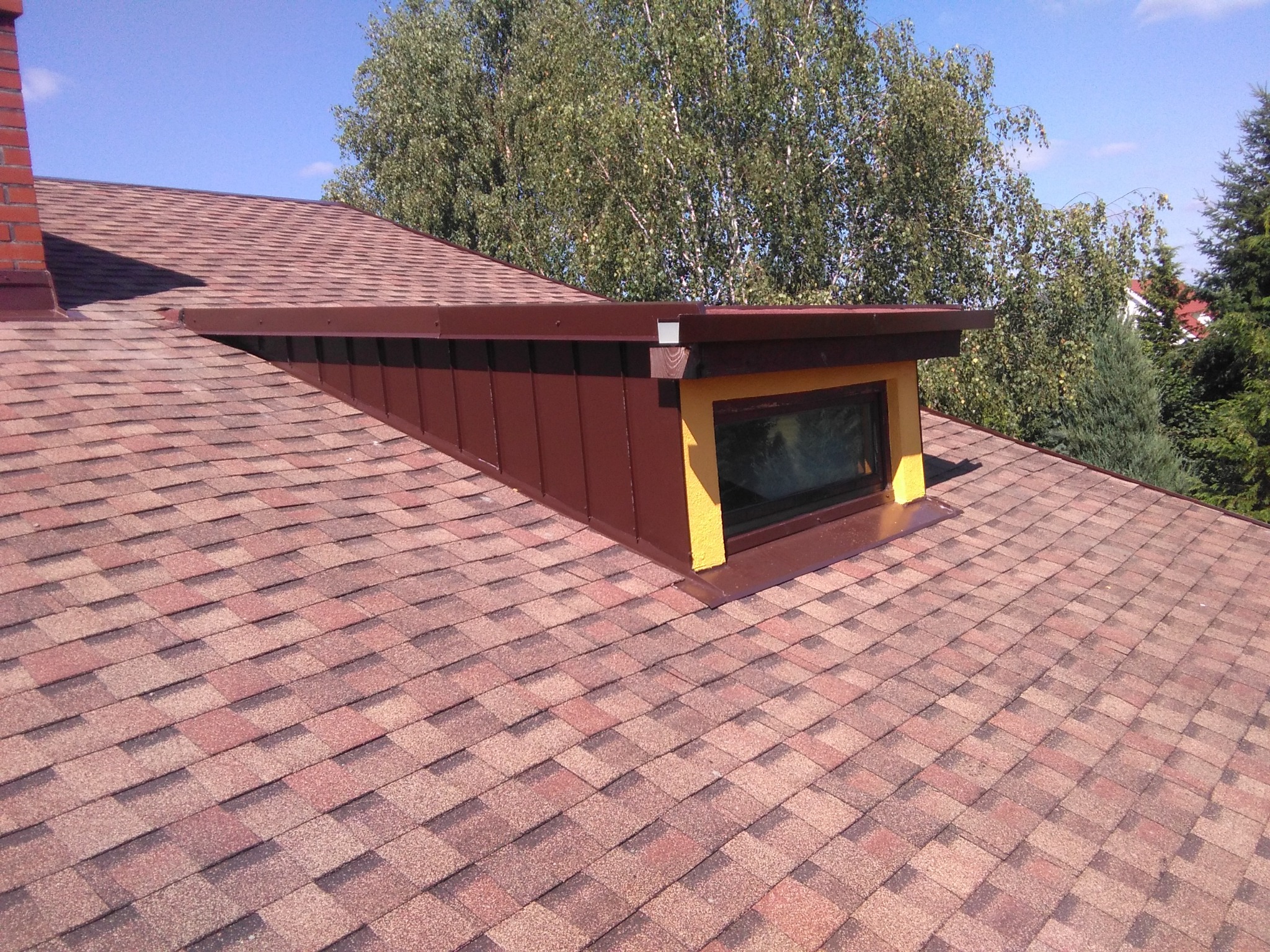 Dach pokryty gontem bitumicznym w odcieniach czerwieni i brązu, z oknem dachowym obudowanym brązową blachą i żółtymi elementami wykończeniowymi, widok z góry na tle zieleni drzew.