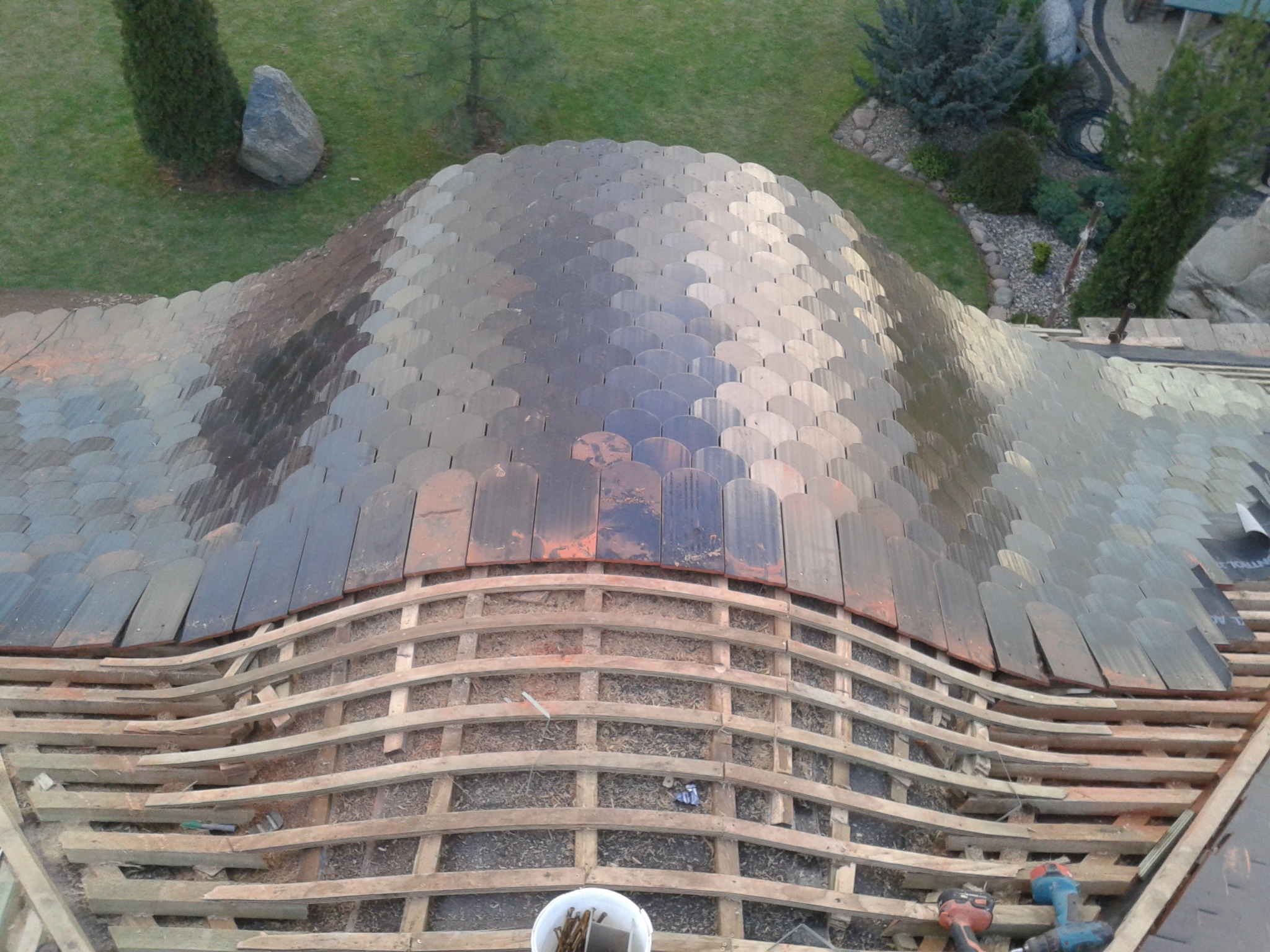 Układanie dachówki łuskowej na drewnianej konstrukcji dachu o nieregularnym kształcie, widok z góry na częściowo pokryty dach, narzędzia pracy w pobliżu.