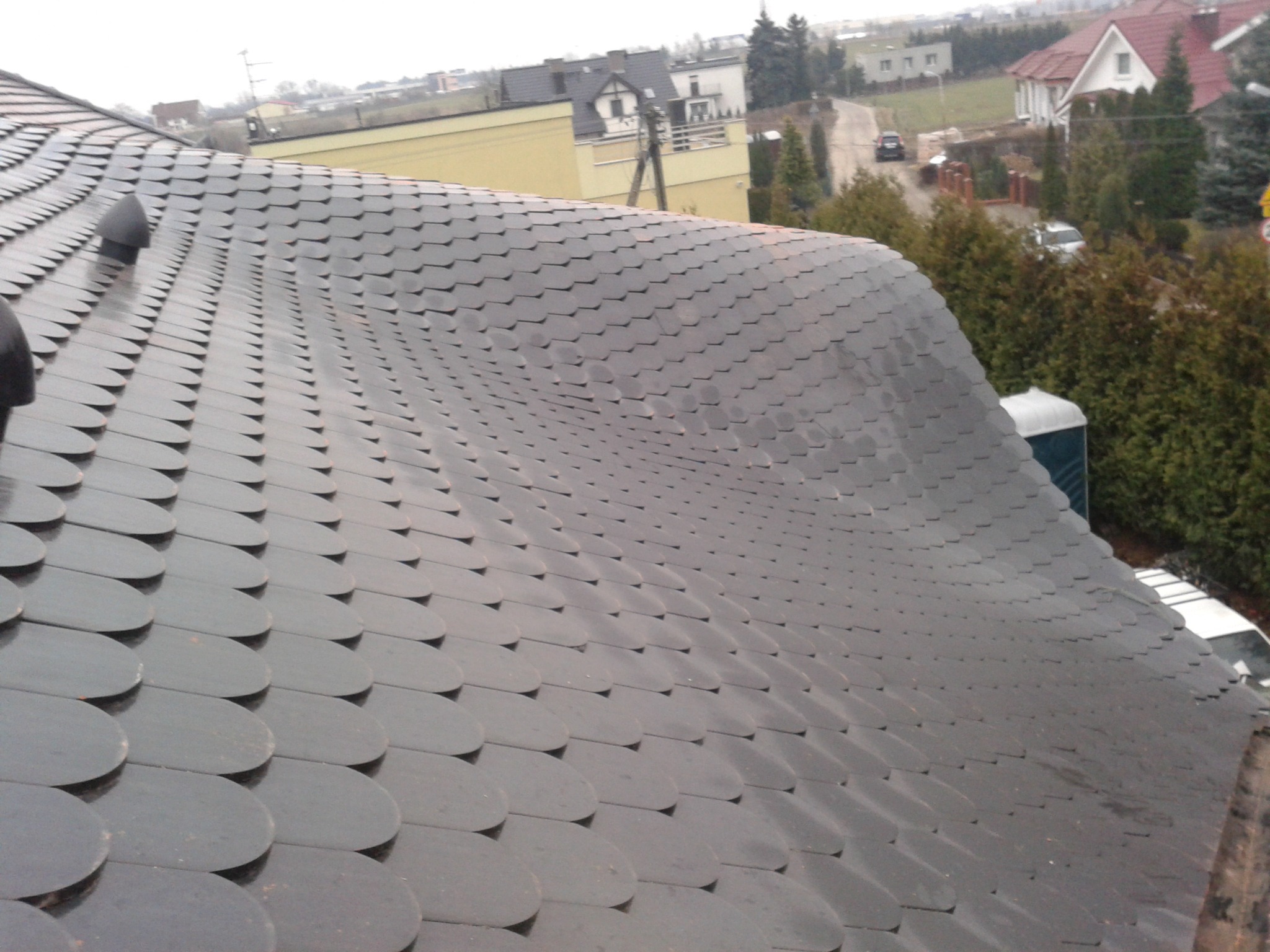Dach pokryty łuską bitumiczną z kominkami wentylacyjnymi, widok z góry na tle zabudowań i zieleni