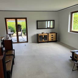 Igor Yarmolitskyi - Jasne, przestronne wnętrze pokoju z drewnianymi meblami i dużymi oknami wychodzącymi na ogród. Świeżo wykończona podłoga z jasnych płytek. Widoczny fragment systemu audio z pilotem na półce.