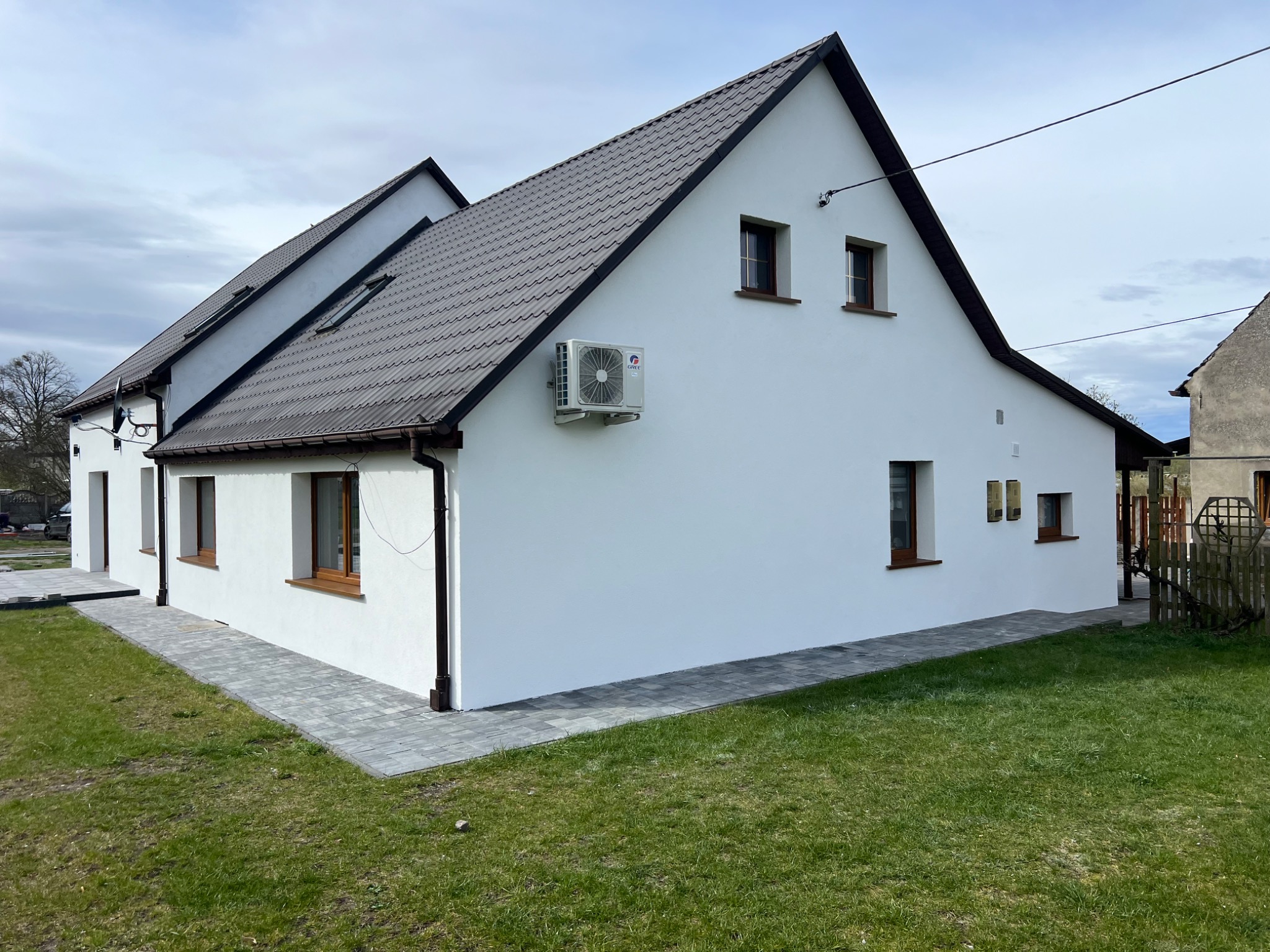 Odnowiony dom jednorodzinny z białą elewacją, brązowym dachem i oknami, z klimatyzatorem na ścianie i nową kostką brukową wokół budynku.