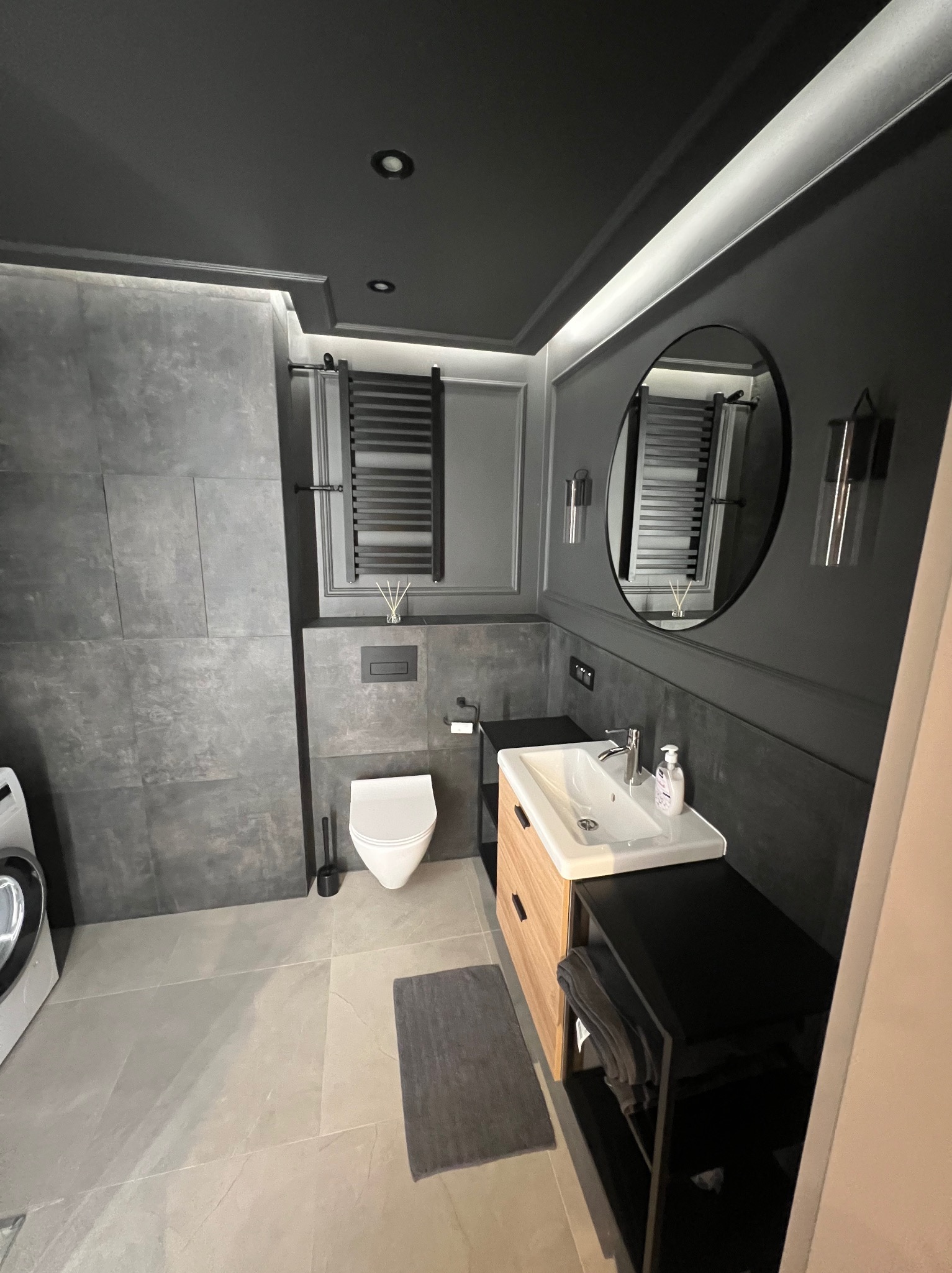 Nowoczesna łazienka w odcieniach szarości z drewnianą szafką pod umywalką, okrągłym lustrem i grzejnikiem drabinkowym, podświetlona listwą LED na suficie.