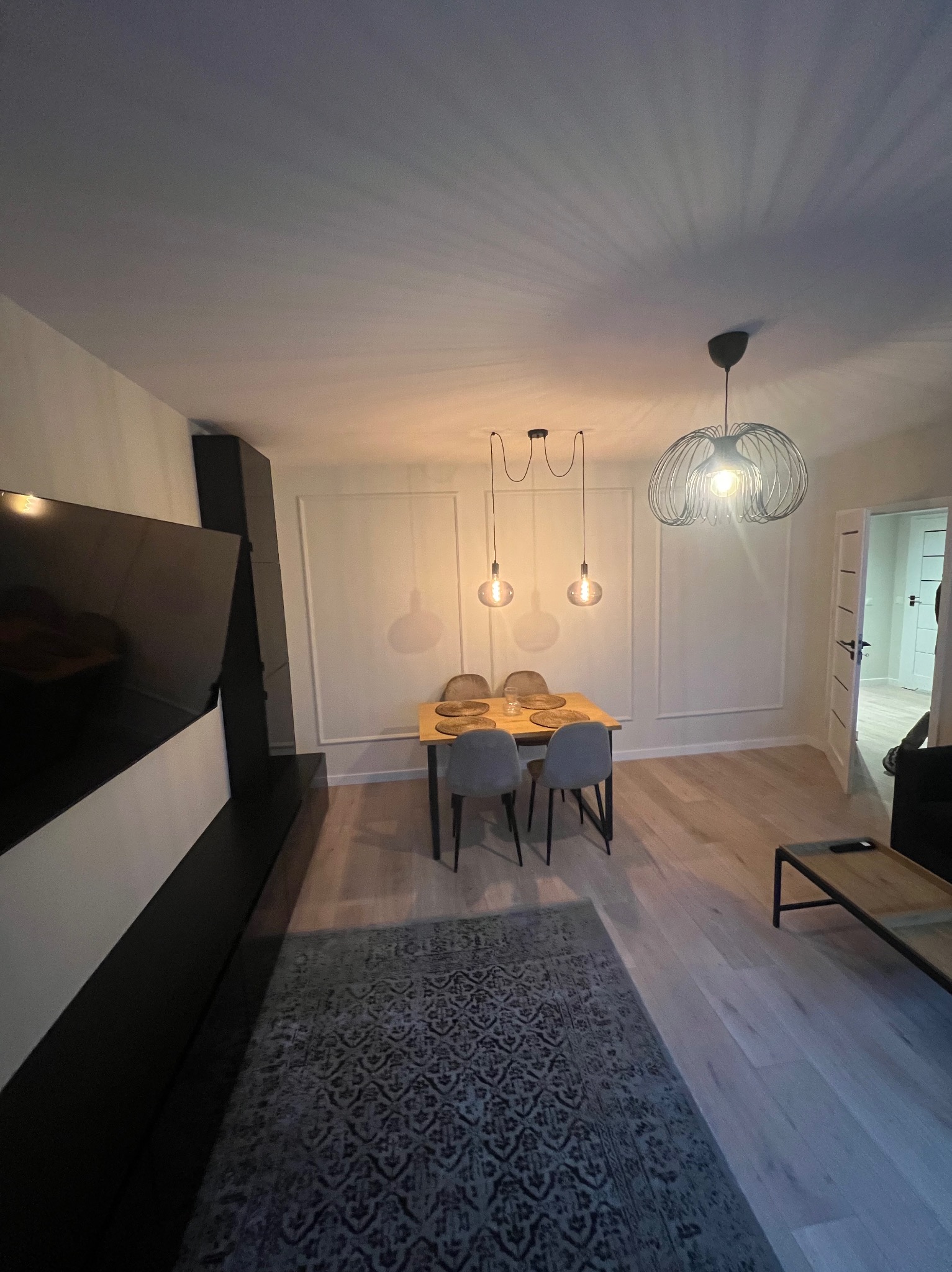 Nowoczesne wnętrze: stół jadalny z czterema krzesłami, oświetlony wiszącymi lampami, dywan z wzorem, parkiet, dekoracyjne panele ścienne i czarny element zabudowy TV.