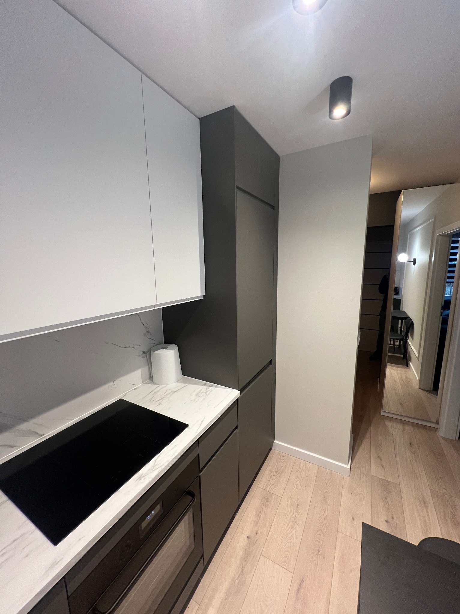 Nowoczesna kuchnia w odcieniach bieli i szarości, z marmurowym blatem, płytą indukcyjną i widocznym fragmentem piekarnika. W tle widoczny fragment korytarza z lustrem.