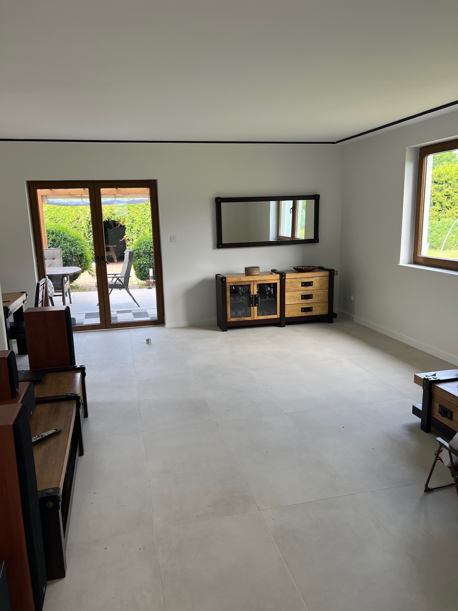 Jasne, przestronne wnętrze pokoju z drewnianymi meblami i dużymi oknami wychodzącymi na ogród. Świeżo wykończona podłoga z jasnych płytek. Widoczny fragment systemu audio z pilotem na półce.