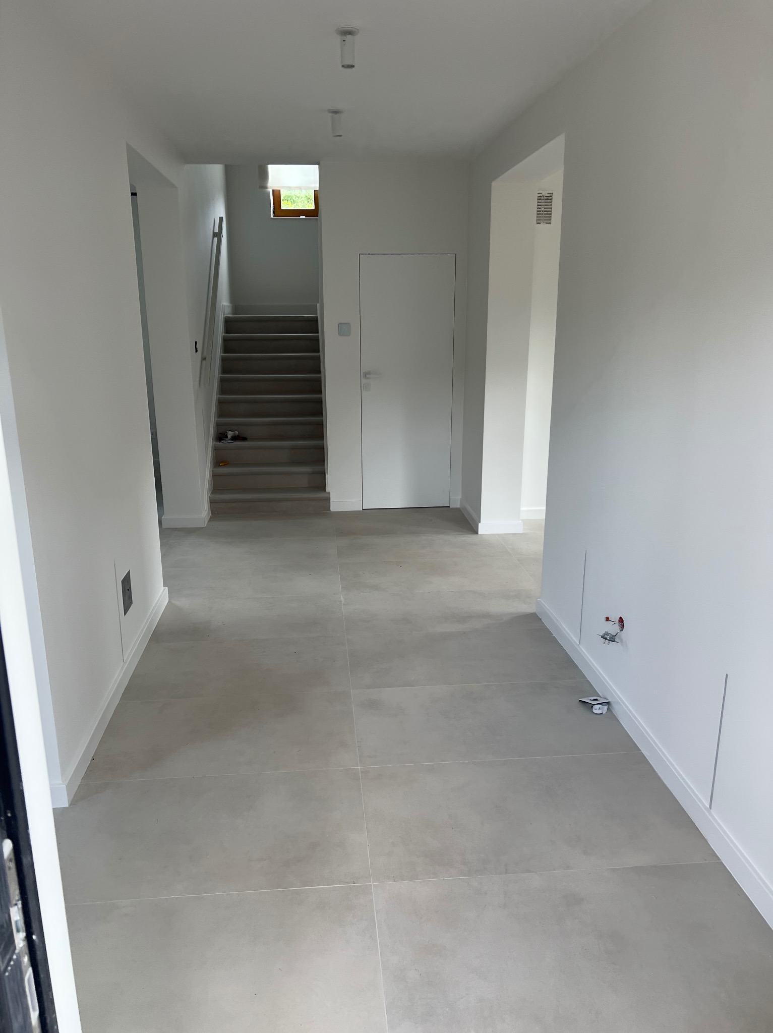 Jasne, minimalistyczne wnętrze domu z szarymi płytkami podłogowymi, białymi ścianami i schodami prowadzącymi na górę. Widoczne instalacje elektryczne i hydrauliczne w ścianie, wskazujące na etap...