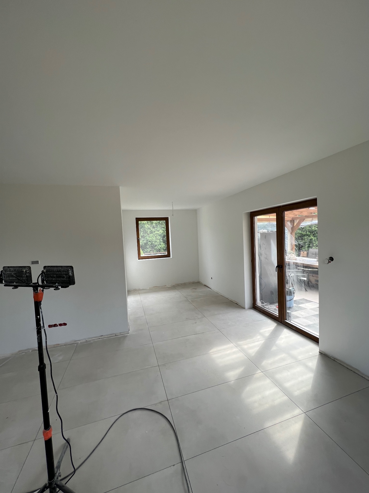 Wnętrze w trakcie prac wykończeniowych: białe ściany po gładziowaniu, szare płytki podłogowe, widoczne okno i drzwi balkonowe z drewnianą ramą, lampa budowlana na statywie.