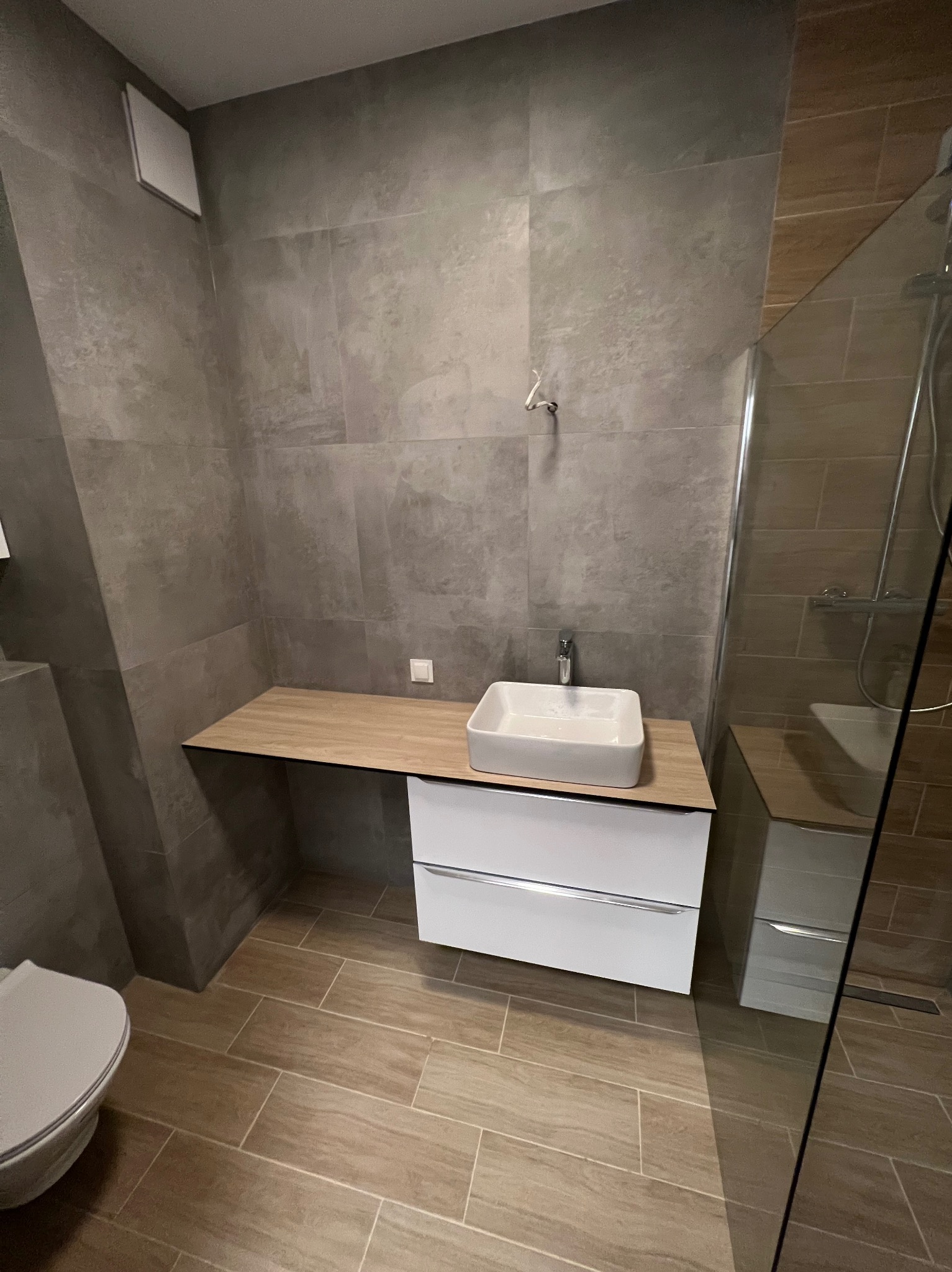Nowoczesna łazienka z płytkami imitującymi beton na ścianach, drewnopodobną podłogą, białą szafką pod umywalką i kabiną prysznicową ze szklaną ścianką.