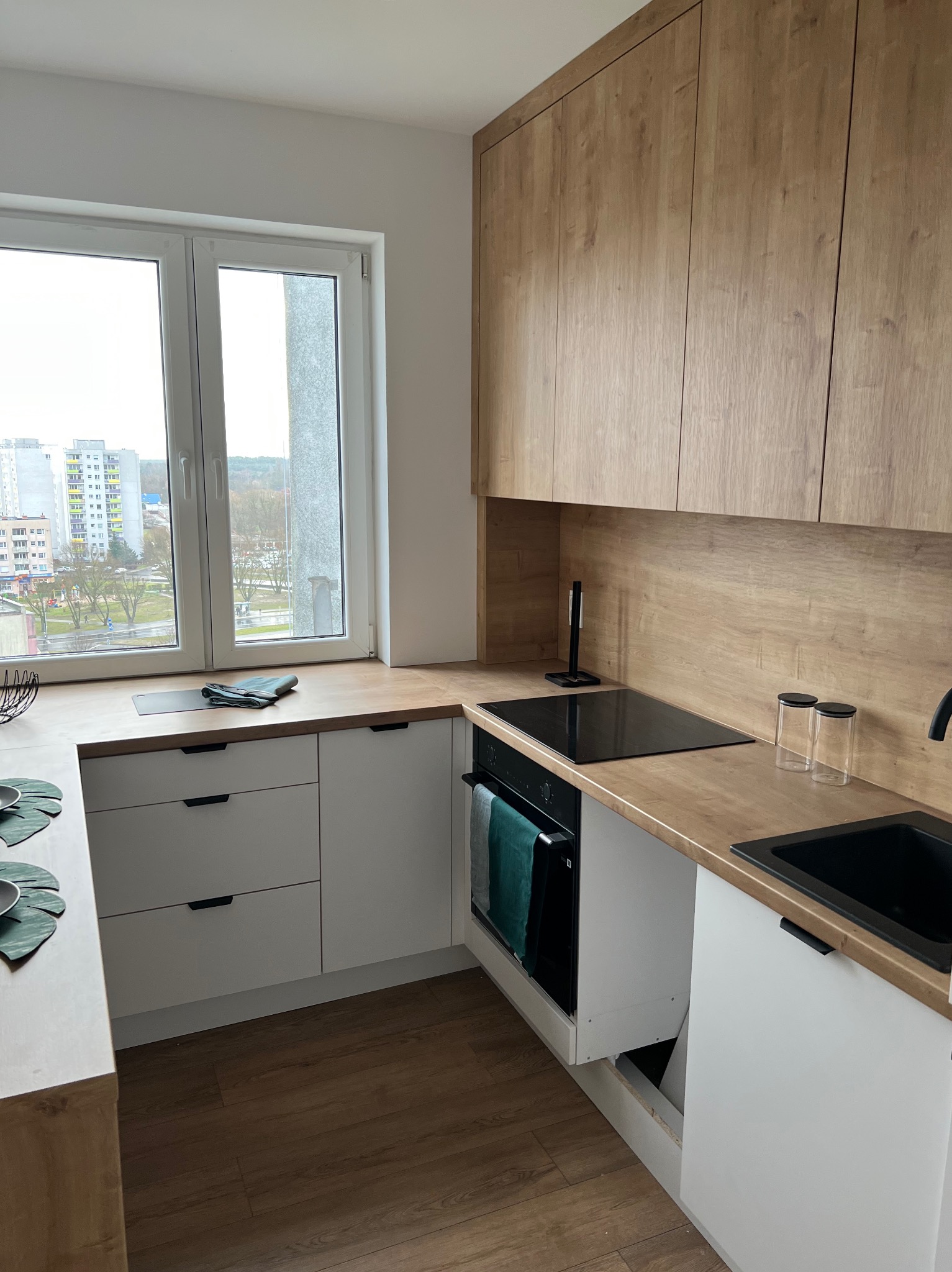 Nowoczesna kuchnia z drewnianymi szafkami wiszącymi i białymi szafkami dolnymi z czarnymi uchwytami, czarnym zlewem i płytą indukcyjną, widok na miasto przez okno.