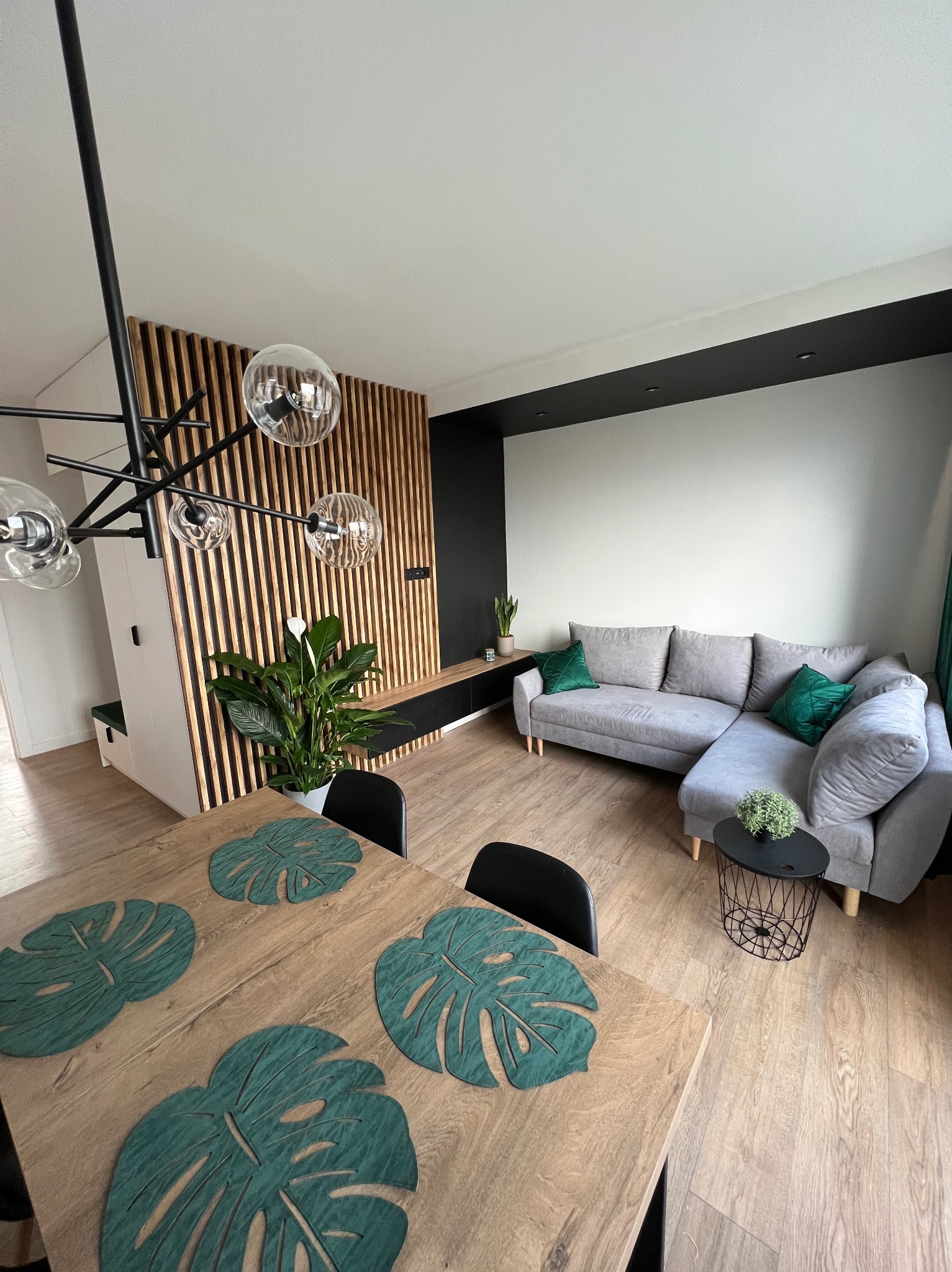 Nowoczesny salon z drewnianą ścianką działową, szarą sofą z zielonymi poduszkami, stołem z podkładkami w kształcie liści monstery i designerską lampą wiszącą.