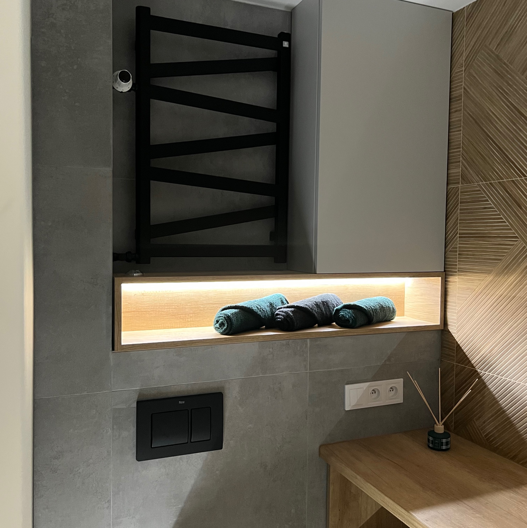 Nowoczesna łazienka z szarymi płytkami, podświetlaną półką z ręcznikami w odcieniach zieleni i czarnym grzejnikiem drabinkowym, akcentowana drewnianą półką i dekoracyjną ścianą z wzorzystych płytek.