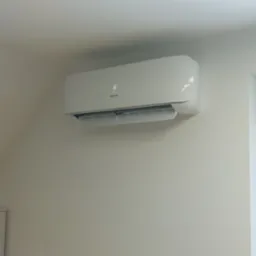 Biały klimatyzator Hisense z wyświetlaczem temperatury 16 stopni Celsjusza, zamontowany na białej ścianie w pomieszczeniu z oknem w tle.