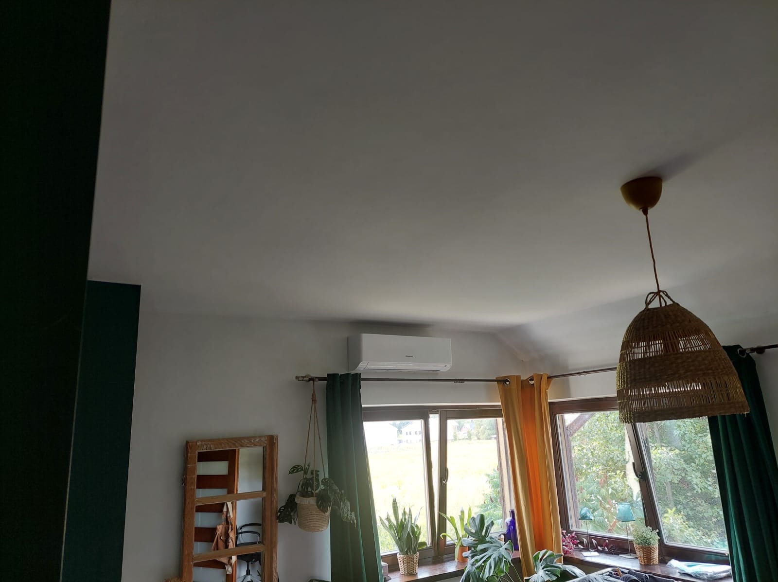 Biały klimatyzator ścienny zamontowany nad oknem z zielonymi i żółtymi zasłonami w jasnym pokoju z drewnianym lustrem i roślinami doniczkowymi.