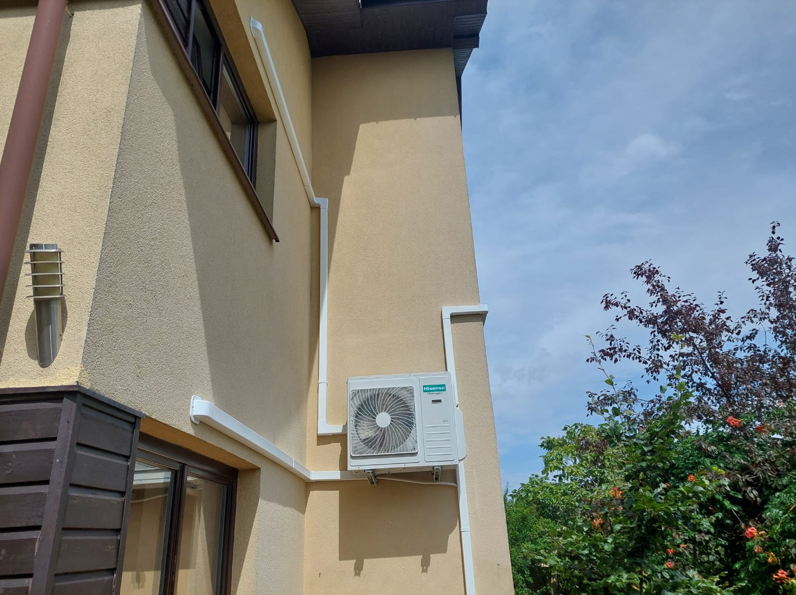 Zewnętrzna jednostka klimatyzacji marki Hisense zamontowana na elewacji domu z widocznymi białymi rurami instalacyjnymi, na tle błękitnego nieba i zieleni drzew.