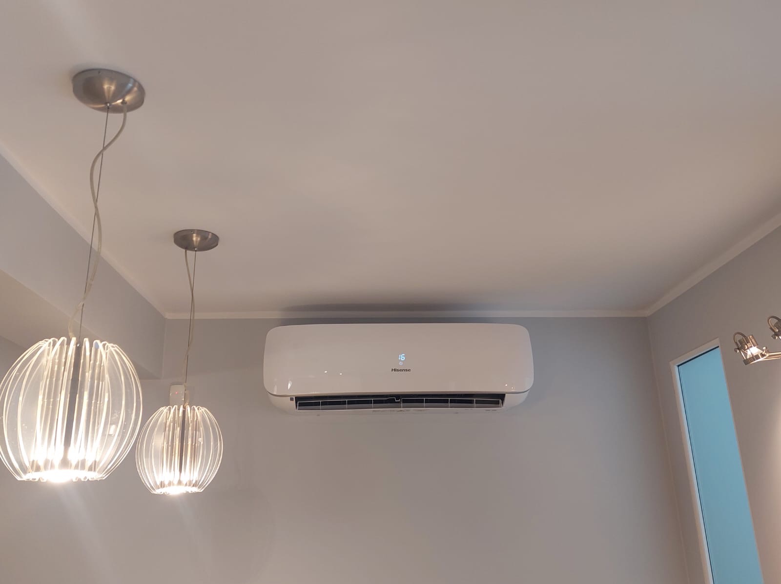 Biały klimatyzator Hisense zamontowany na szarej ścianie, widoczne dwie designerskie lampy wiszące z metalowymi elementami.