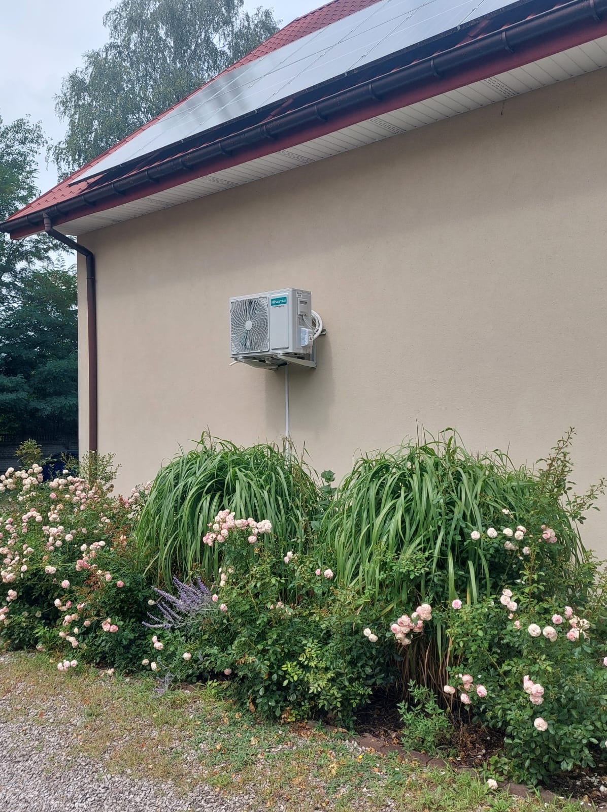 Zewnętrzna jednostka klimatyzacji Hisense zamontowana na beżowej ścianie domu z czerwonym dachem, otoczona bujną roślinnością i różami.