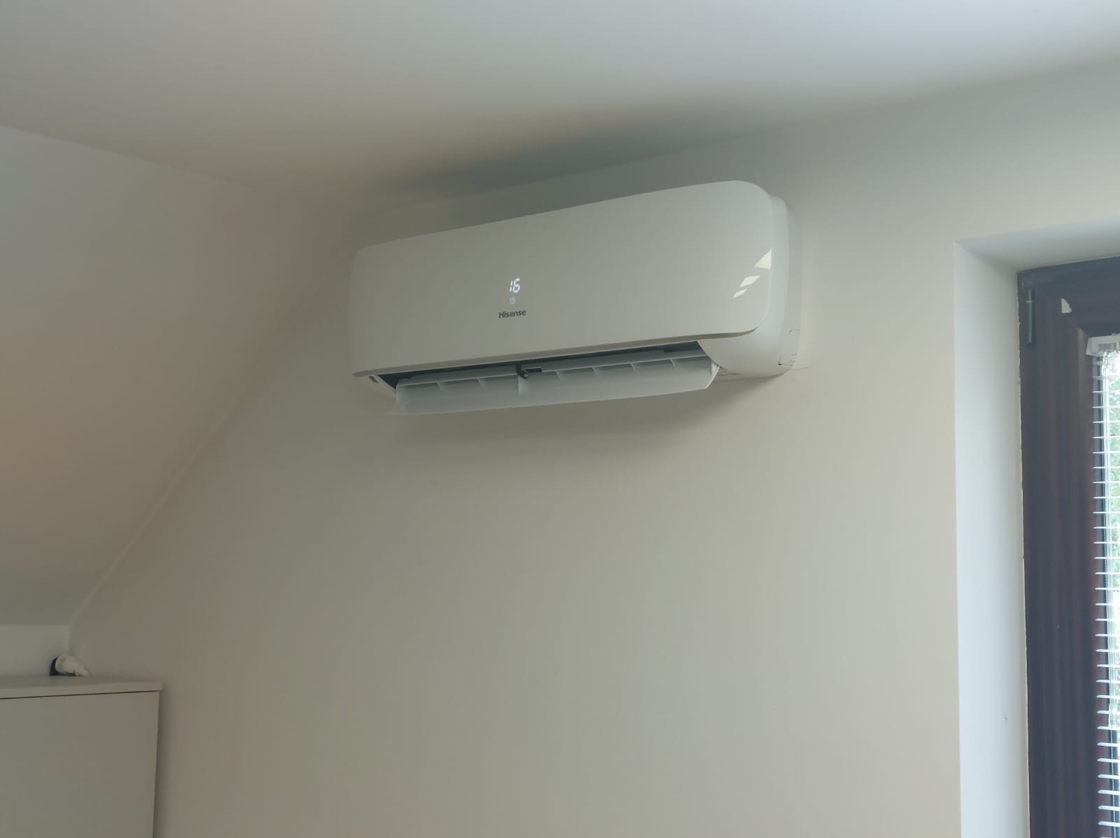 Biały klimatyzator Hisense z wyświetlaczem temperatury 16 stopni Celsjusza, zamontowany na białej ścianie w pomieszczeniu z oknem w tle.