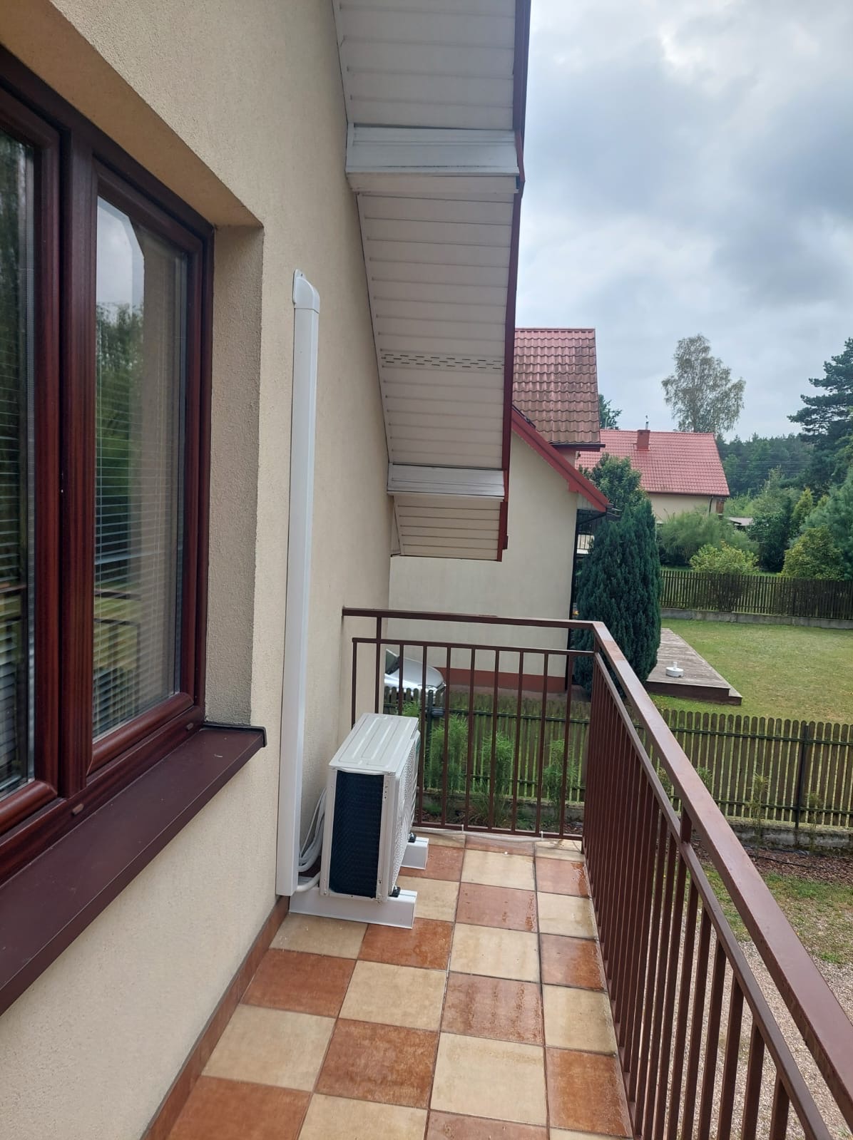 Zewnętrzna jednostka klimatyzacji zamontowana na balkonie domu jednorodzinnego, widoczna brązowa balustrada, okno z brązową ramą i fragment elewacji w kolorze beżowym.