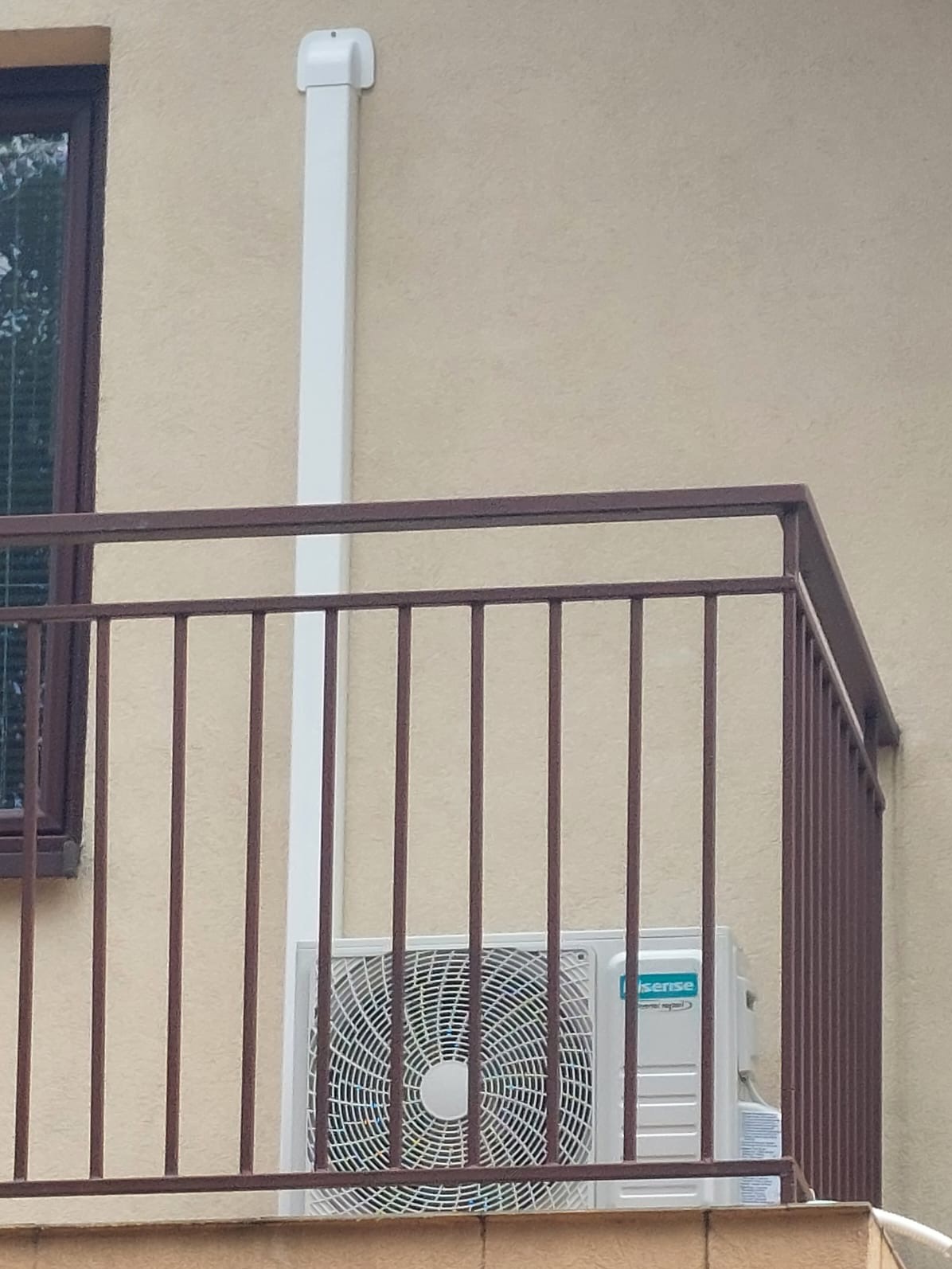 Jednostka zewnętrzna klimatyzacji Hisense zamontowana na balkonie, widoczna przez brązową balustradę, z białą rurą odprowadzającą skropliny.