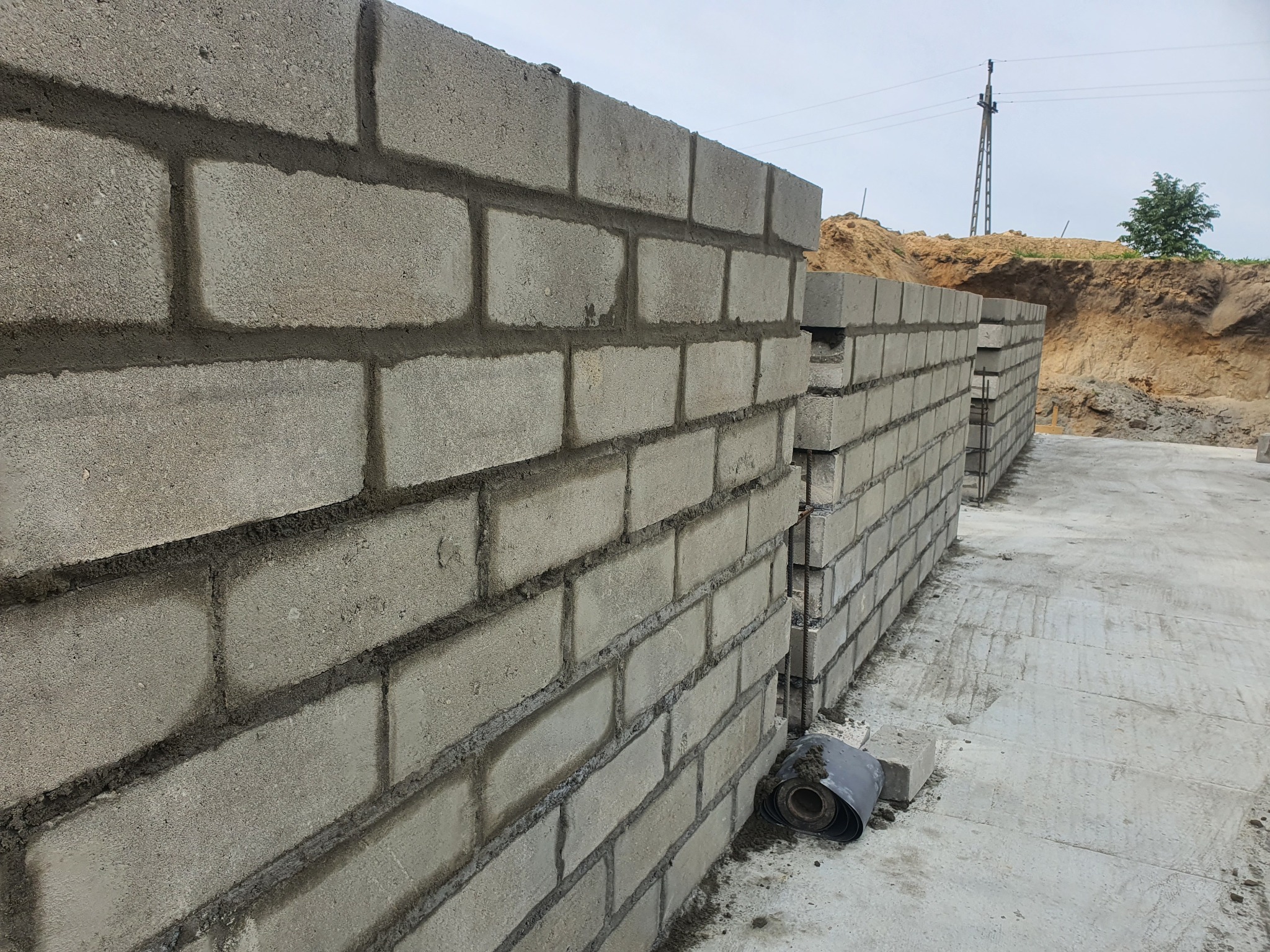 Wznoszenie szarej ściany z bloczków betonowych z widocznymi spoinami, fragment rolki papy na posadzce, w tle wykop ziemny i słup energetyczny.