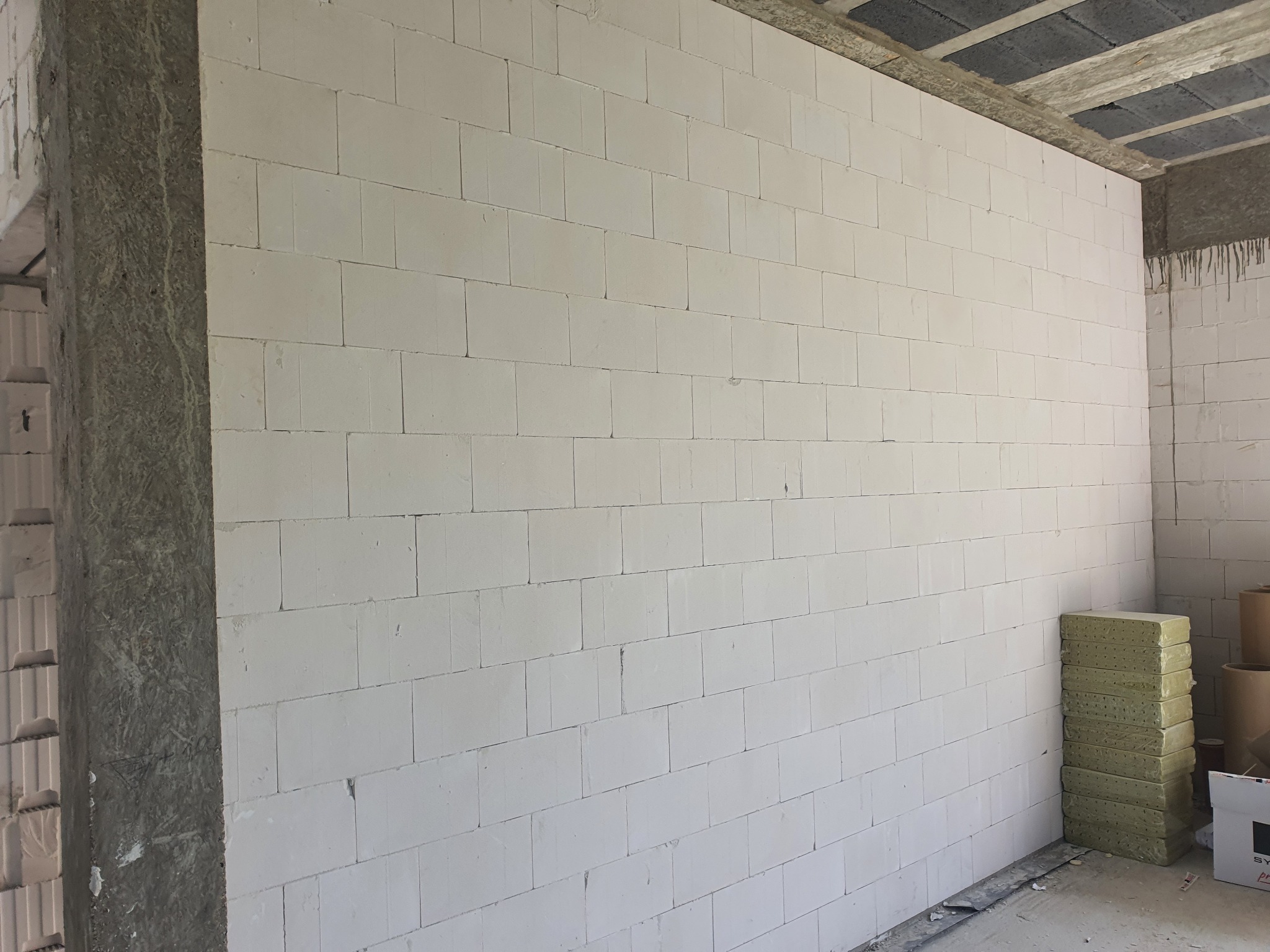 Wnętrze budynku w trakcie budowy: ściana wymurowana z białych bloczków, widoczne zapasy materiału izolacyjnego obok, surowy betonowy strop.