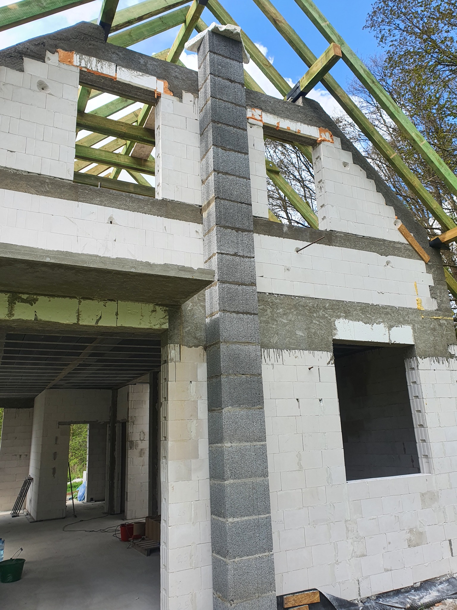 Wznoszenie dwukondygnacyjnej ściany domu z białych bloczków, widoczny fragment komina z szarych bloczków, drewniana konstrukcja dachu i surowy betonowy strop.