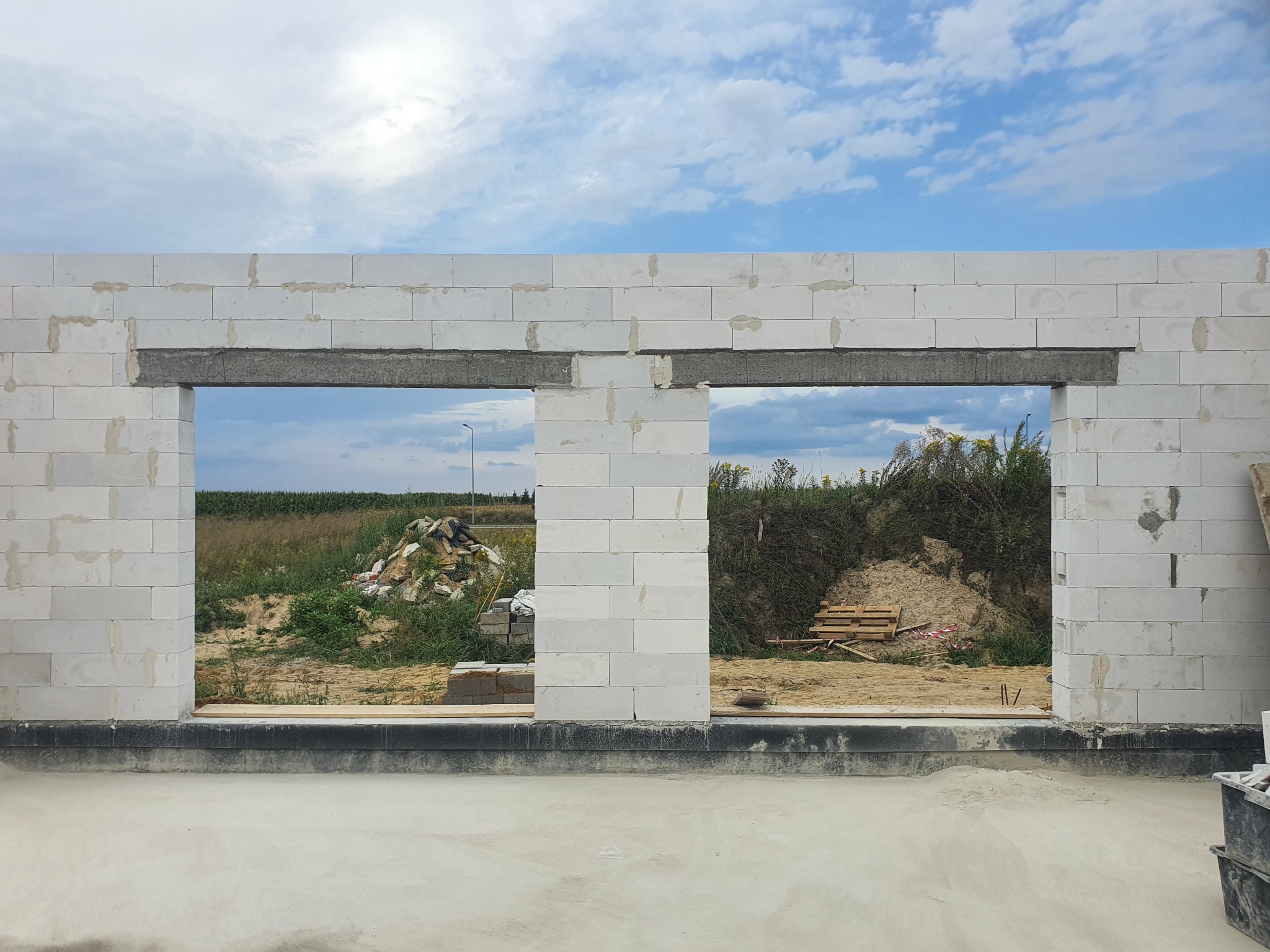 Wznoszenie ścian z białych bloczków betonowych z widocznymi otworami okiennymi, na tle krajobrazu polnego i błękitnego nieba z chmurami.