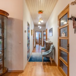 Klaudia Burek The Wonders Agencja Nieruchomości - Jasny, przestronny korytarz z drewnianą podłogą, przeszkloną witryną z dekoracyjnymi przedmiotami, niebieskim dywanem i krzesłem, oraz ozdobnym wieszakiem na ścianie.