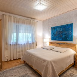 Klaudia Burek The Wonders Agencja Nieruchomości - Jasna sypialnia z drewnianymi meblami, wzorzystą tapetą i turkusowym kilimem nad łóżkiem