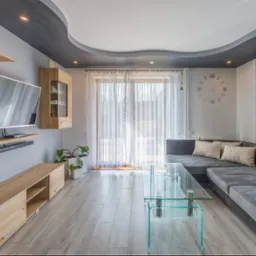 Jasne wnętrze salonu z szarym narożnikiem, szklanym stolikiem kawowym, telewizorem na ścianie i dekoracyjnym oświetleniem sufitowym.