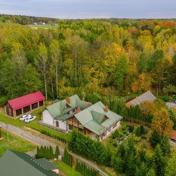 Klaudia Burek The Wonders Agencja Nieruchomości - Panoramiczny widok z drona na duży dom z zielonym dachem, otoczony jesiennym lasem i zabudowaniami gospodarczymi. Dwa białe samochody zaparkowane obok czerwonego garażu.