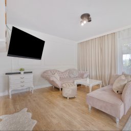 Klaudia Burek The Wonders Agencja Nieruchomości - Jasne wnętrze z pudrowo-różowymi meblami w stylu glamour, drewnianą podłogą i kominkiem. Telewizor na ścianie. Okno z zasłonami w tle.