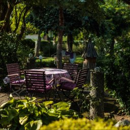 Klaudia Burek The Wonders Agencja Nieruchomości - Urokliwy zakątek ogrodu z drewnianym zestawem mebli ogrodowych w odcieniach różu, otoczony bujną zielenią i dekoracyjnymi elementami. Idealne miejsce na relaks.