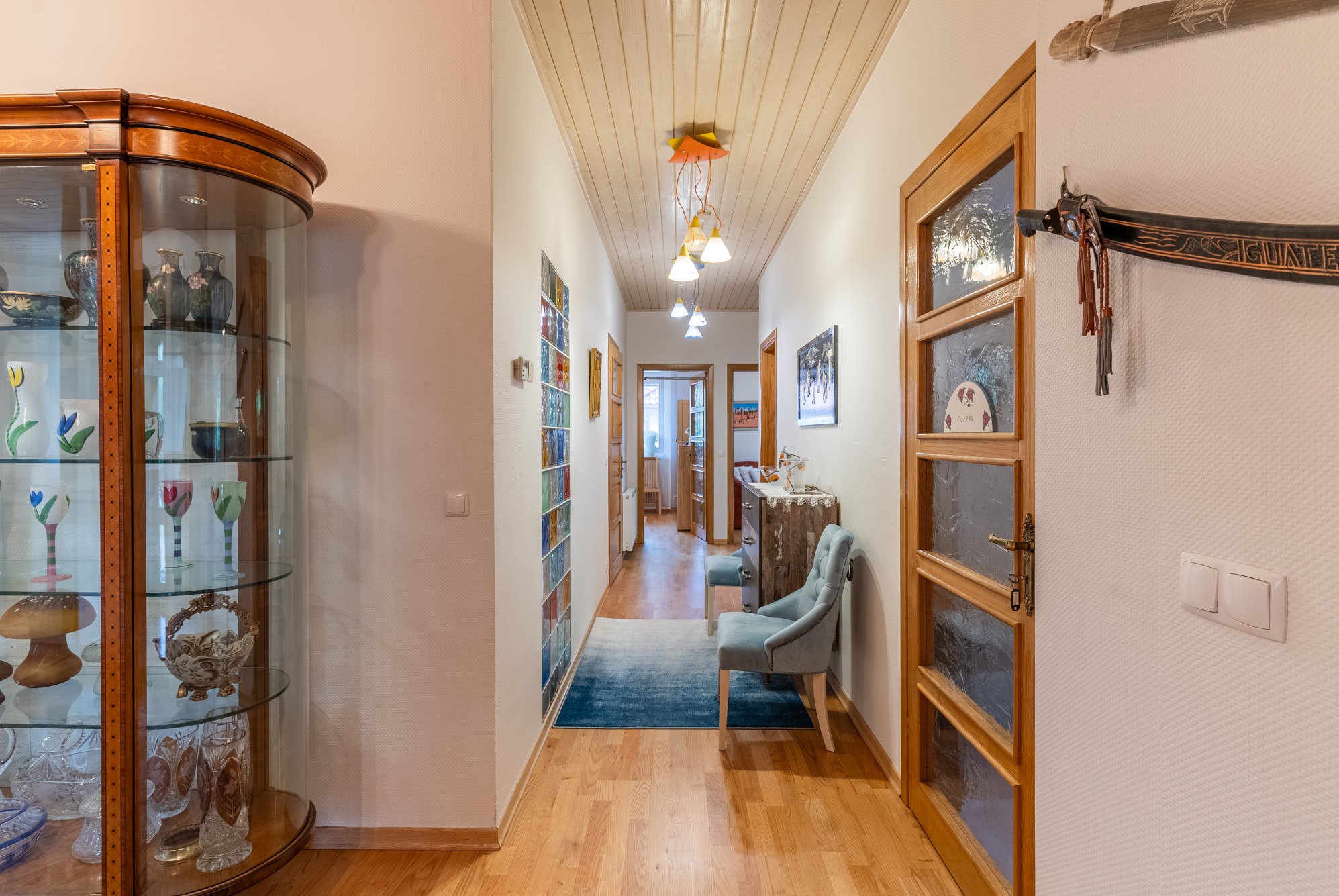 Jasny, przestronny korytarz z drewnianą podłogą, przeszkloną witryną z dekoracyjnymi przedmiotami, niebieskim dywanem i krzesłem, oraz ozdobnym wieszakiem na ścianie.