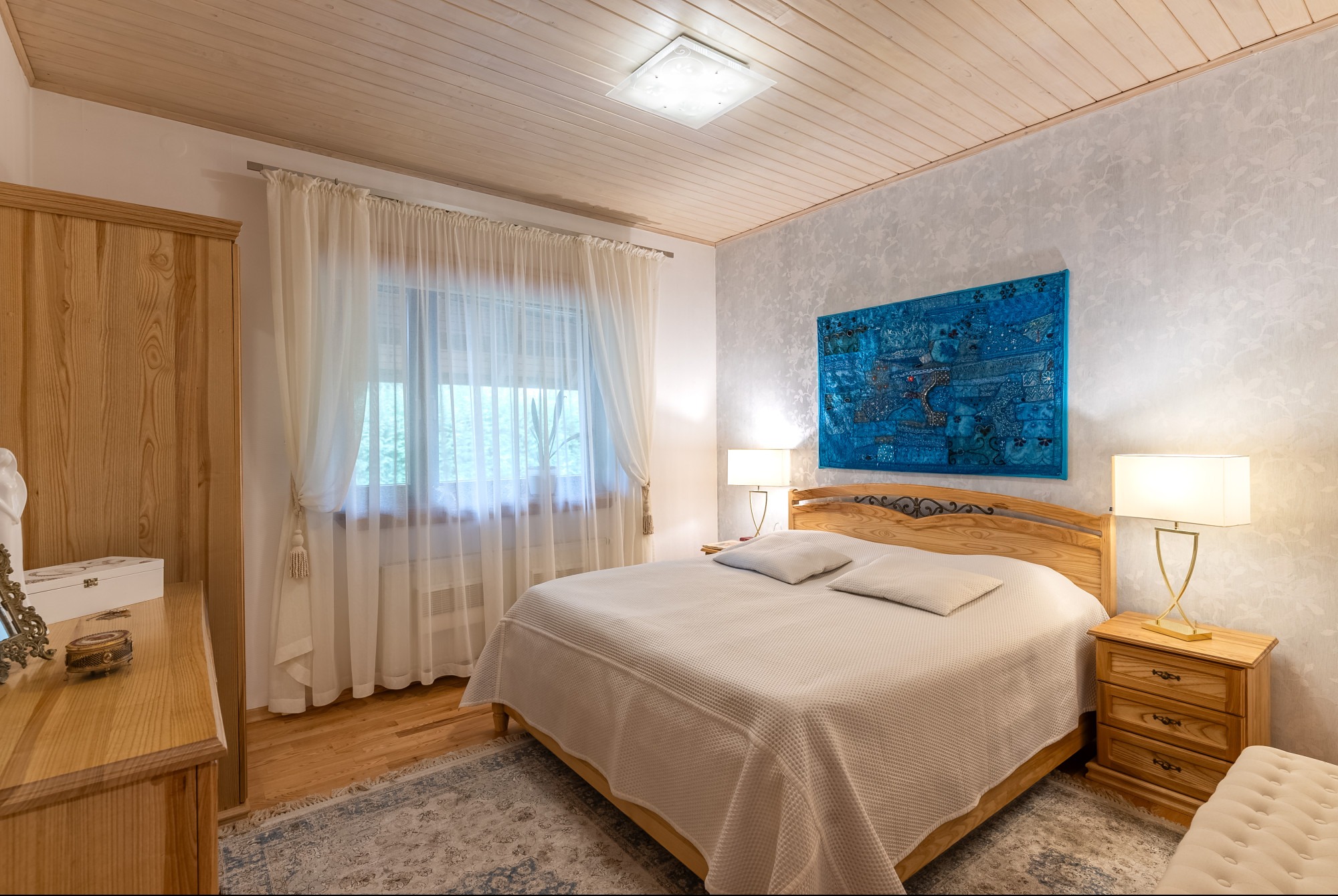 Jasna sypialnia z drewnianymi meblami, wzorzystą tapetą i turkusowym kilimem nad łóżkiem