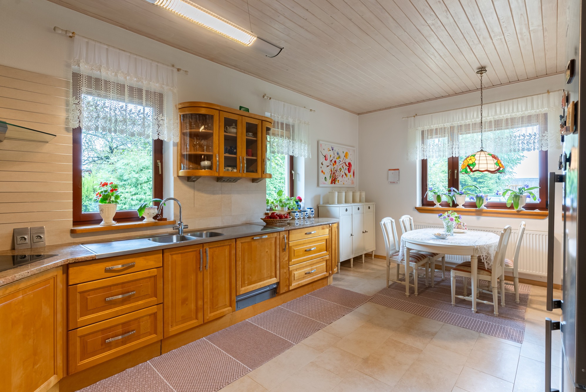 Jasna, przestronna kuchnia z drewnianymi meblami, okrągłym stołem i kwiatami w oknach, utrzymana w stylu retro z nowoczesnymi akcentami.