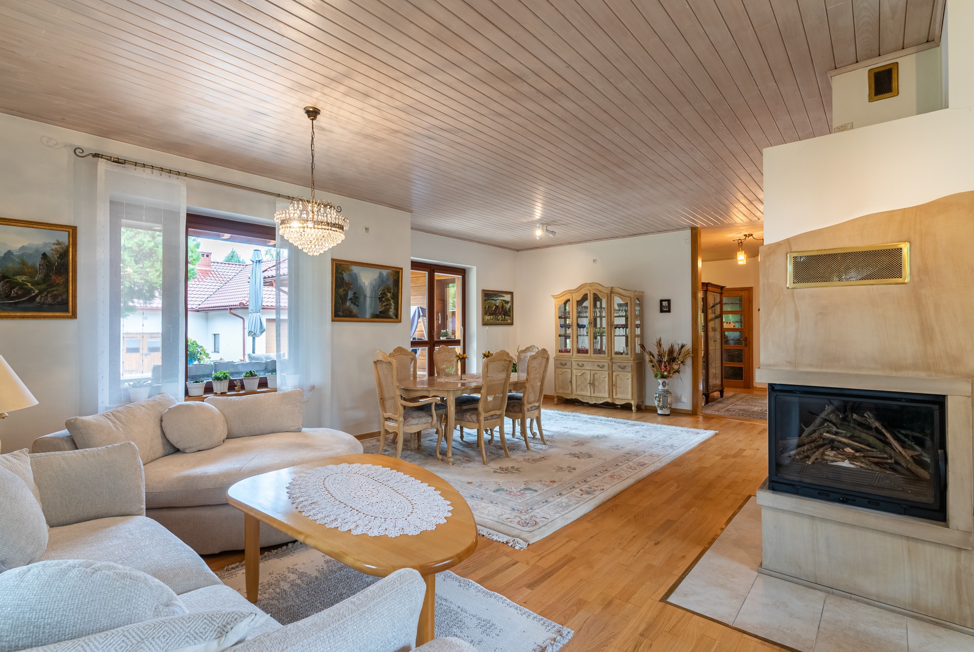 Jasny salon z drewnianą podłogą, kominkiem z obudową w odcieniach beżu, kryształowym żyrandolem i meblami w stylu vintage