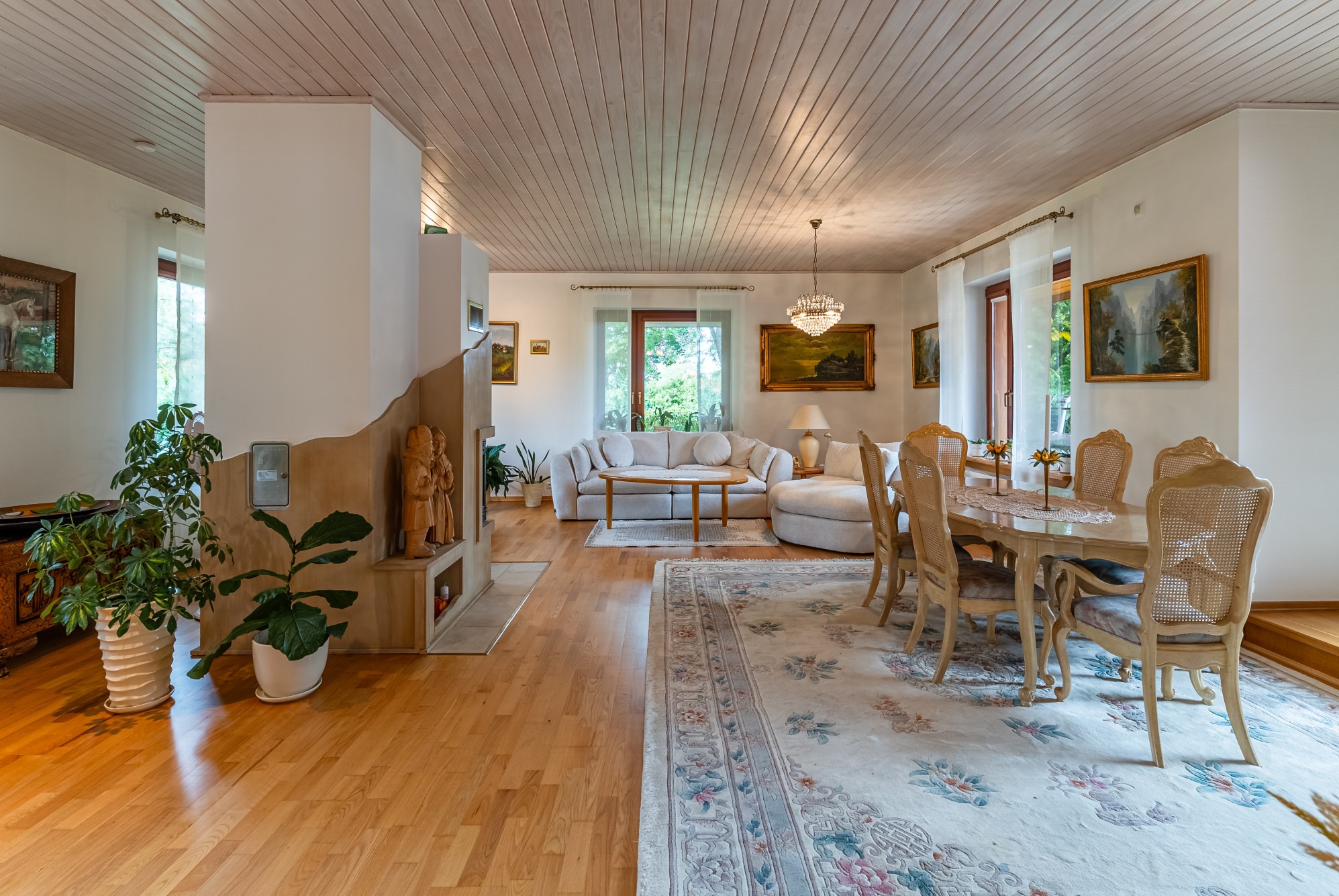 Jasny salon z drewnianą podłogą, beżową sofą, okrągłym stolikiem kawowym, stołem jadalnym z krzesłami z wiklinowymi oparciami i dywanem w kwiatowe wzory, oświetlony kryształowym żyrandolem.