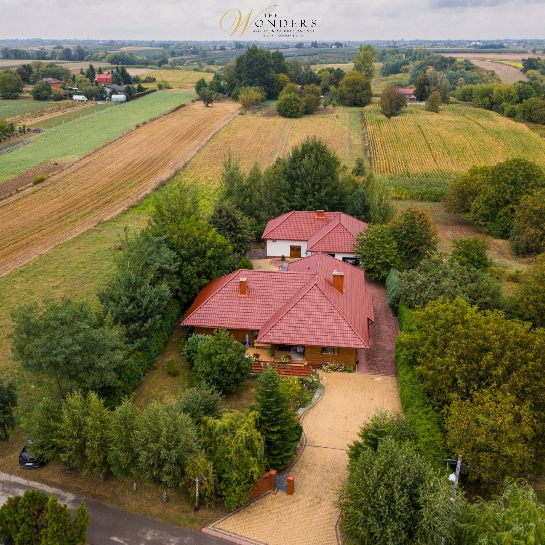 Widok z lotu ptaka na parterowy dom z czerwoną dachówką otoczony zielenią, z polami w tle, w słoneczny dzień.