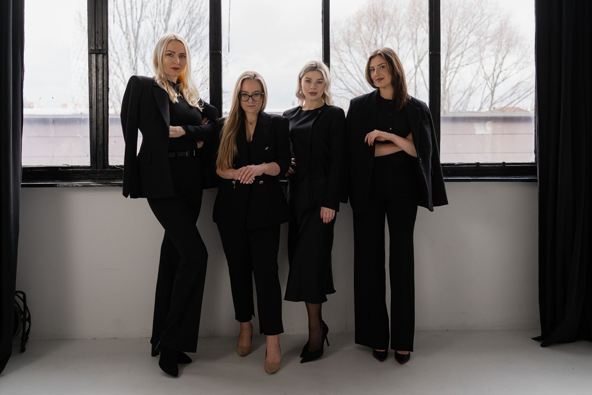 Portret grupowy czterech eleganckich kobiet w czarnych garniturach na tle okna z widokiem na miasto.