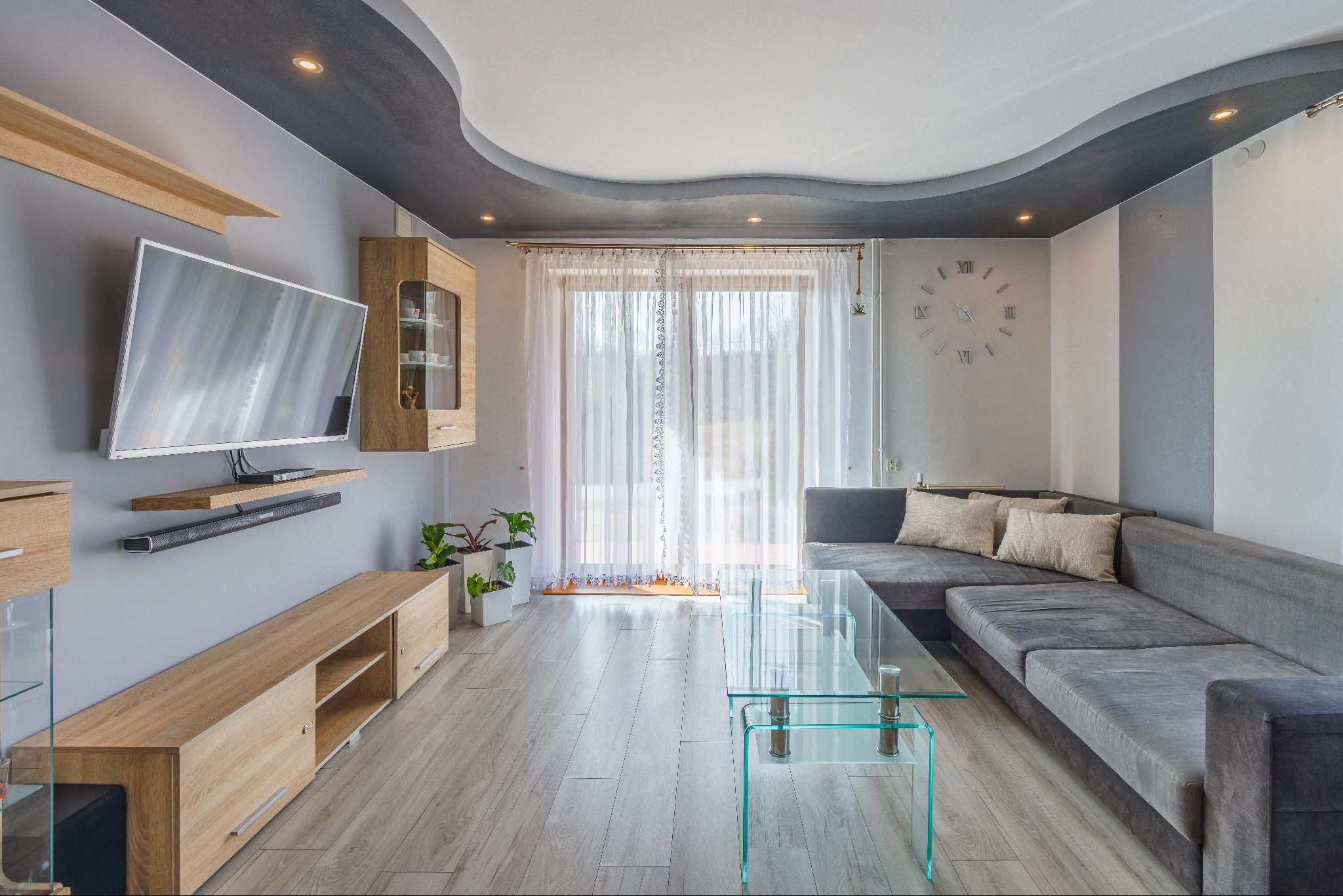 Jasne wnętrze salonu z szarym narożnikiem, szklanym stolikiem kawowym, telewizorem na ścianie i dekoracyjnym oświetleniem sufitowym.