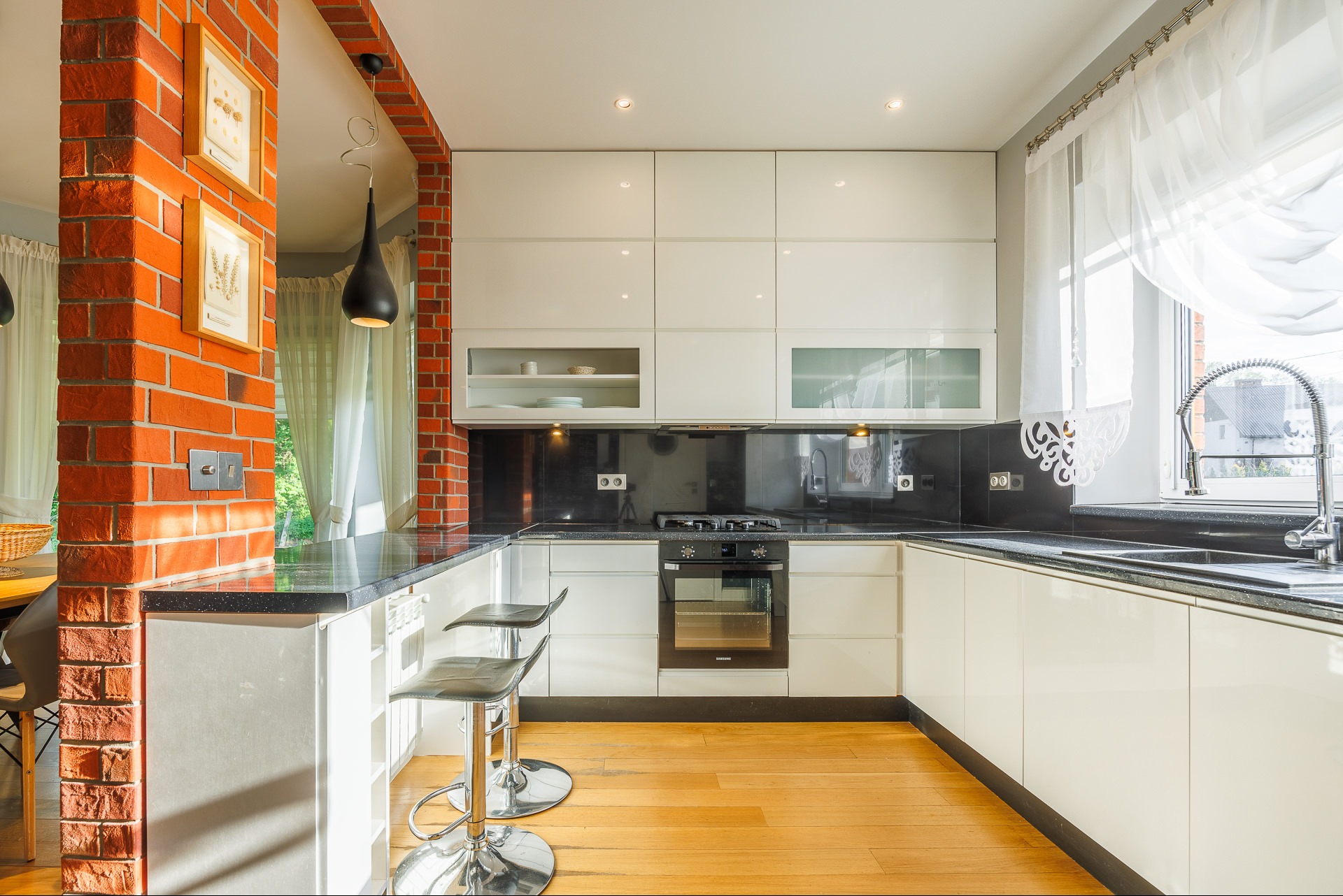 Nowoczesna kuchnia z ceglanym filarem w Rzeszowie. Białe szafki, czarny blat, hoker barowy. Jasne wnętrze z oknem i designerską lampą.