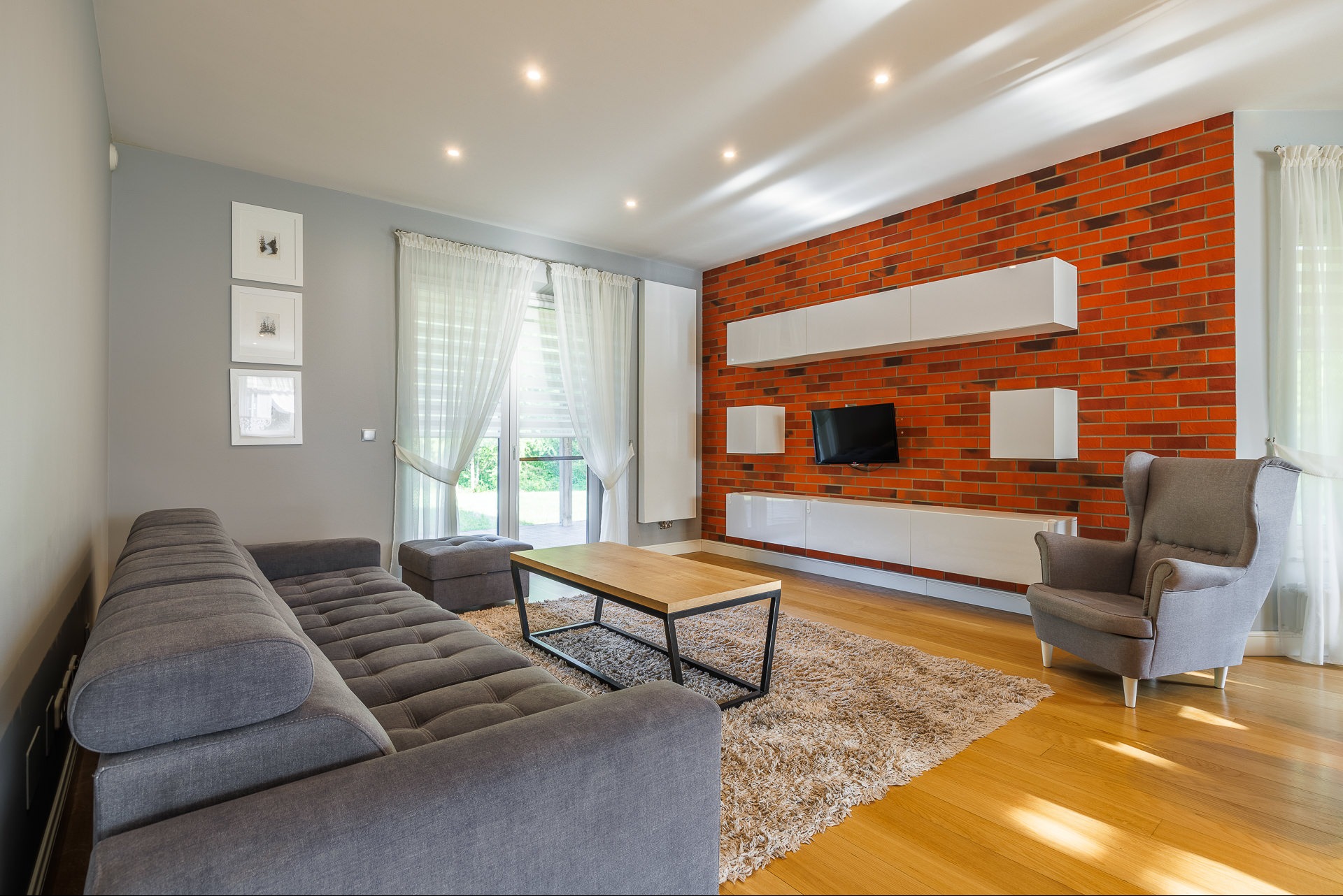 Jasne wnętrze salonu z ceglaną ścianą, szarą sofą i fotelami. Nowoczesne meble, drewniana podłoga i dywan tworzą przytulną przestrzeń. Widok na okno z firanami.